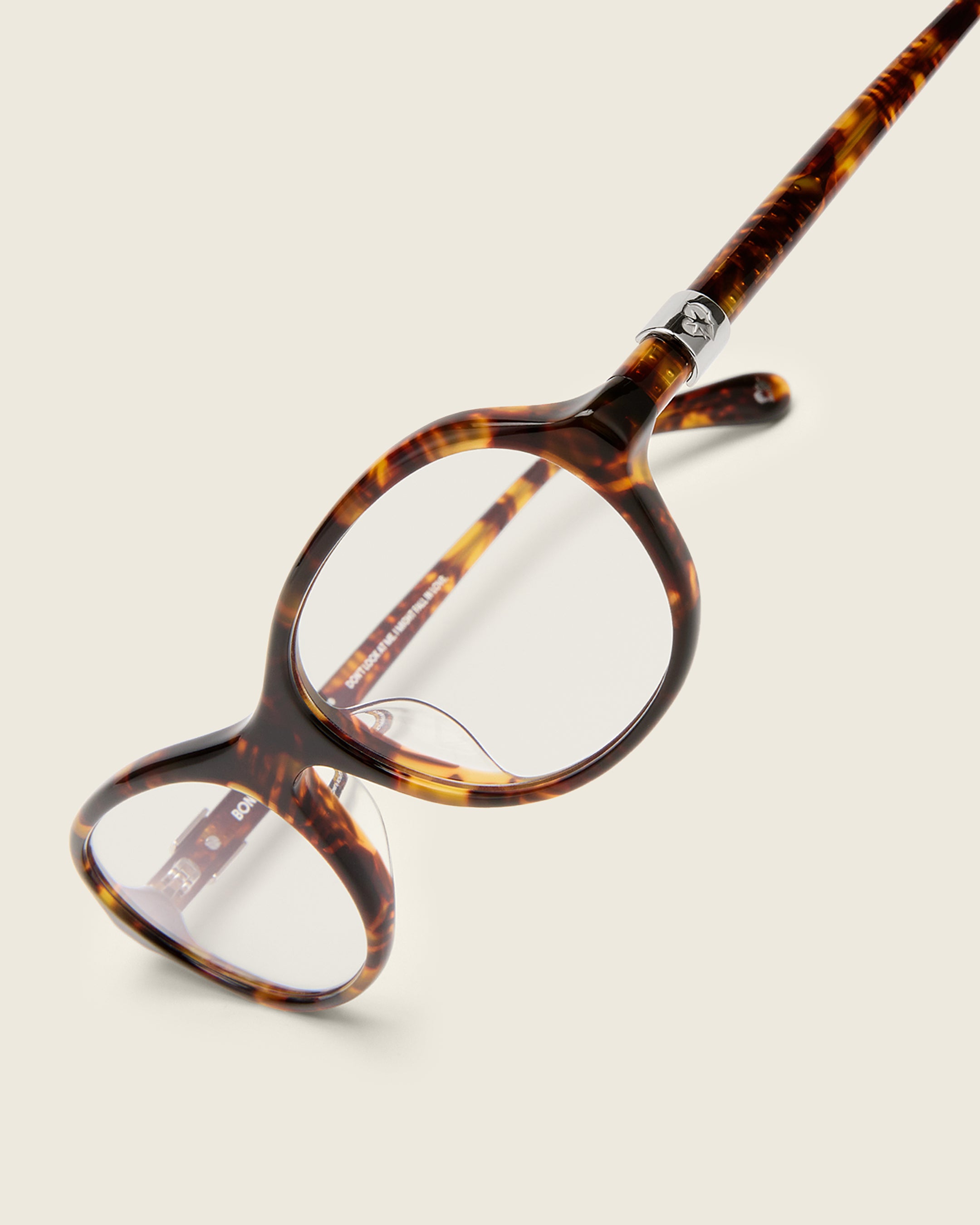 Rounded tortoise acetate frames. Features raised browline curvature on a beige background. Features custom metal heart logo detail on arm temple. Detail view.