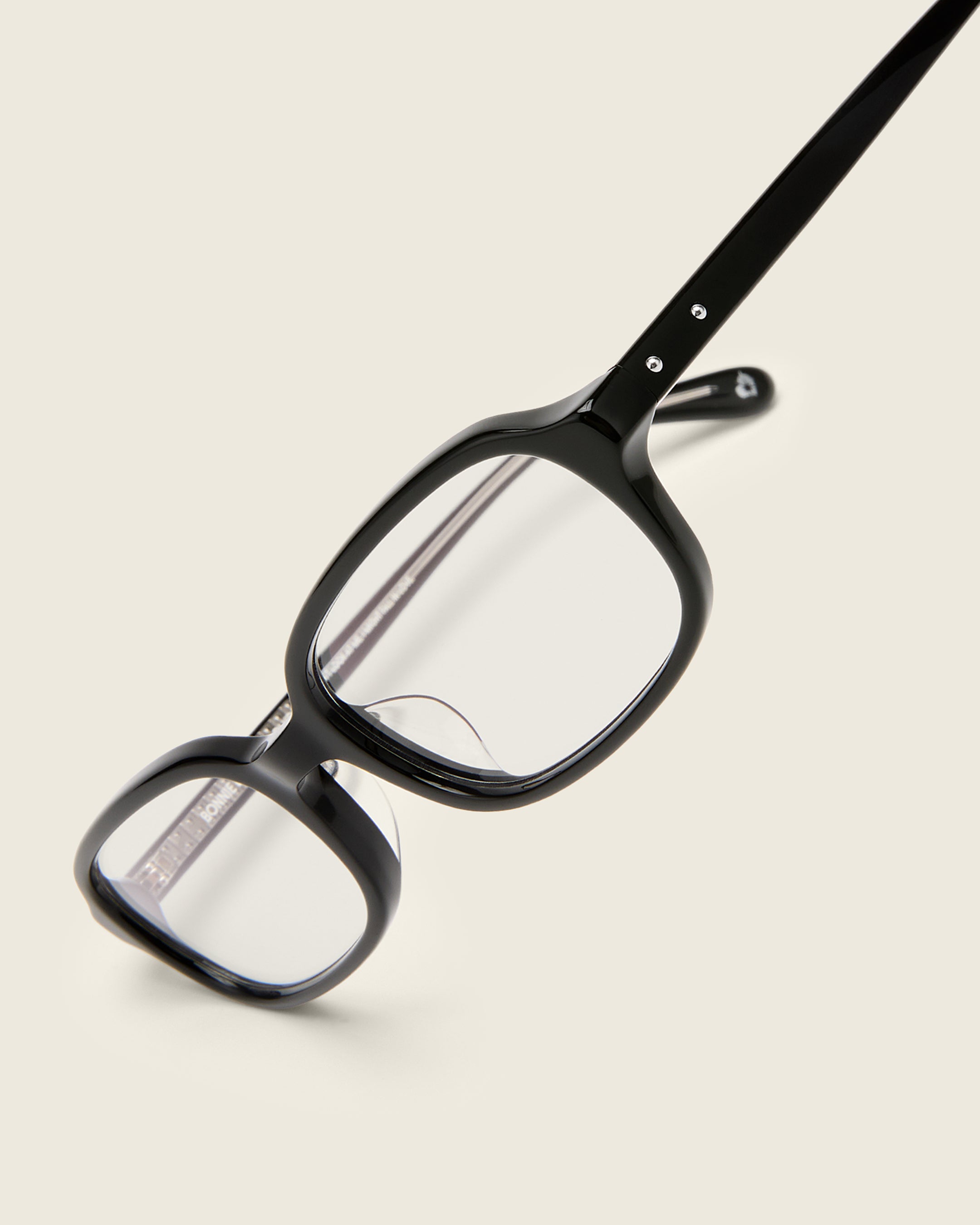 Rectangular black acetate frames with softened corners and a low-profile bridge on a beige background. Detail view.