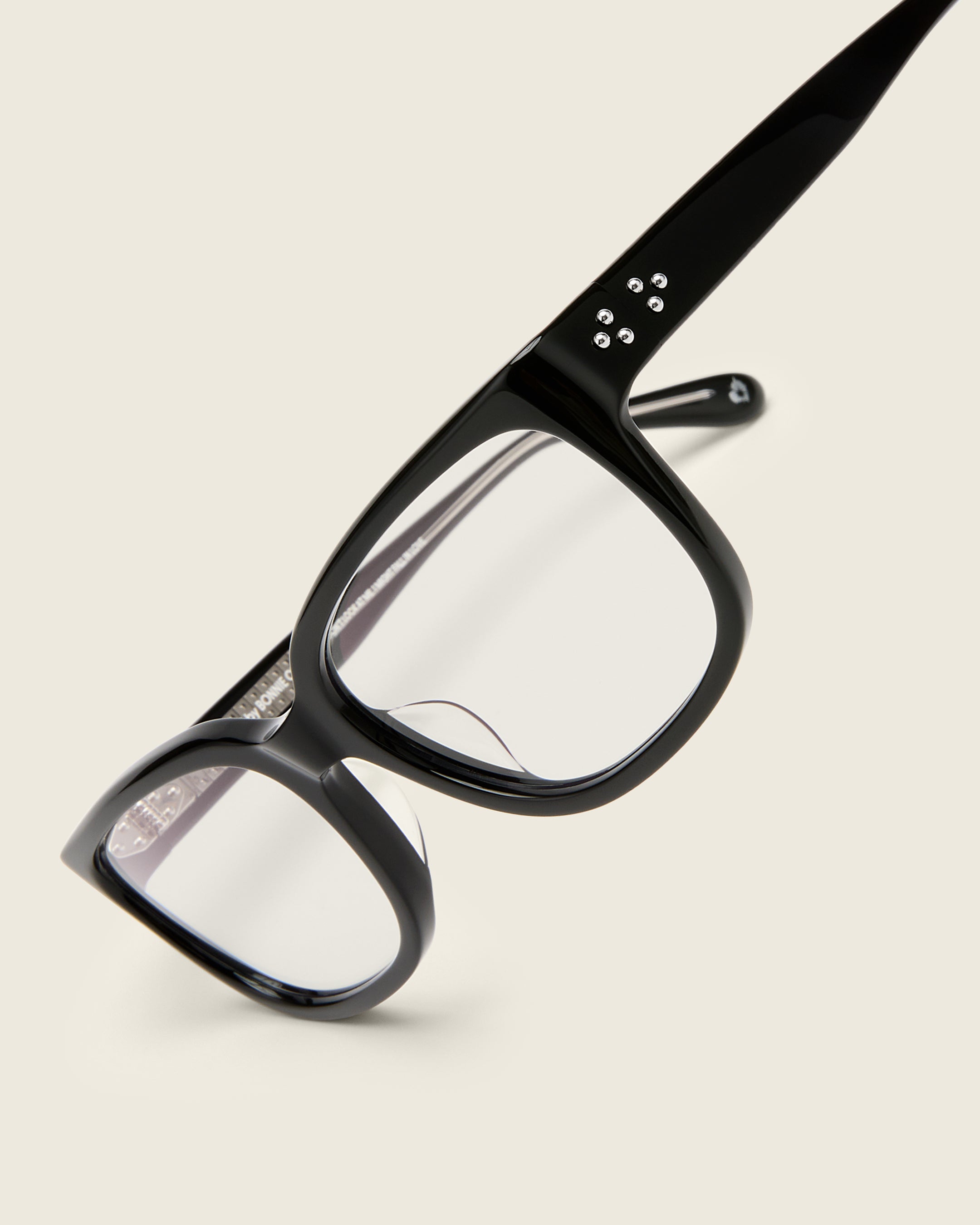 Bold, square-shaped black acetate frames on a beige background. Detail view. Mirrored metal bead detail on arm temples.