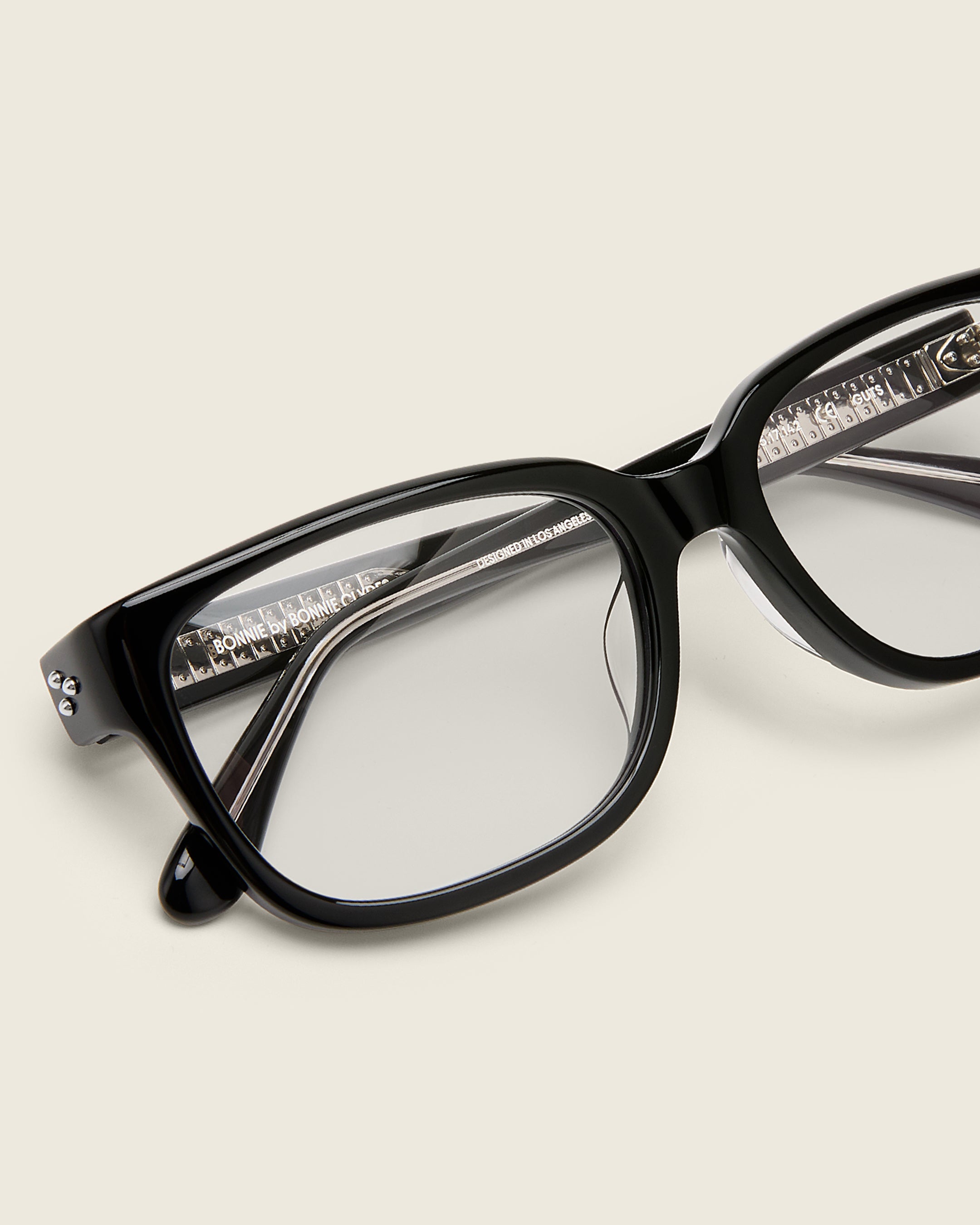 Bold, square-shaped black acetate frames on a beige background. Detail view. Mirrored metal bead detail on arm temples.