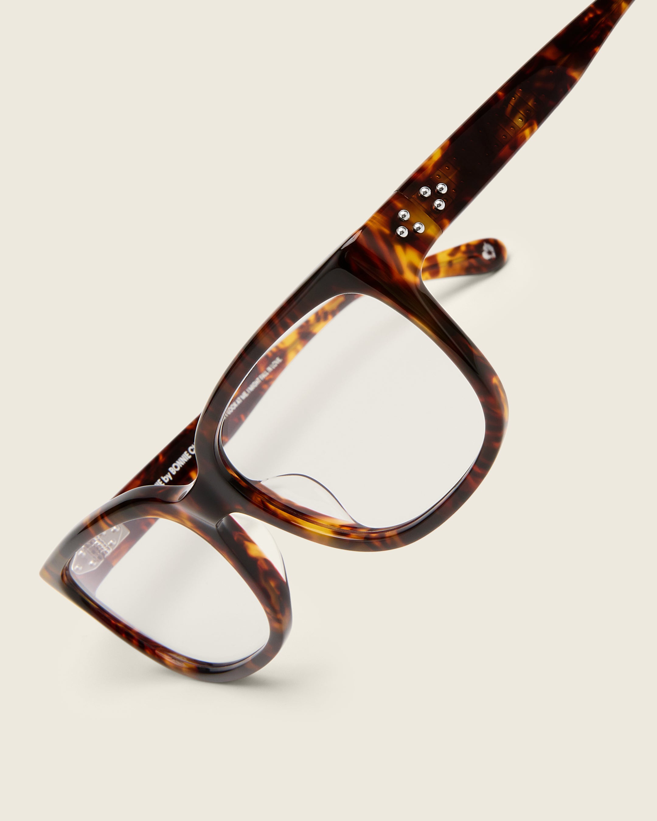 Bold, square-shaped tortoise acetate frames on a beige background. Detail view. Mirrored metal bead detail on arm temples.