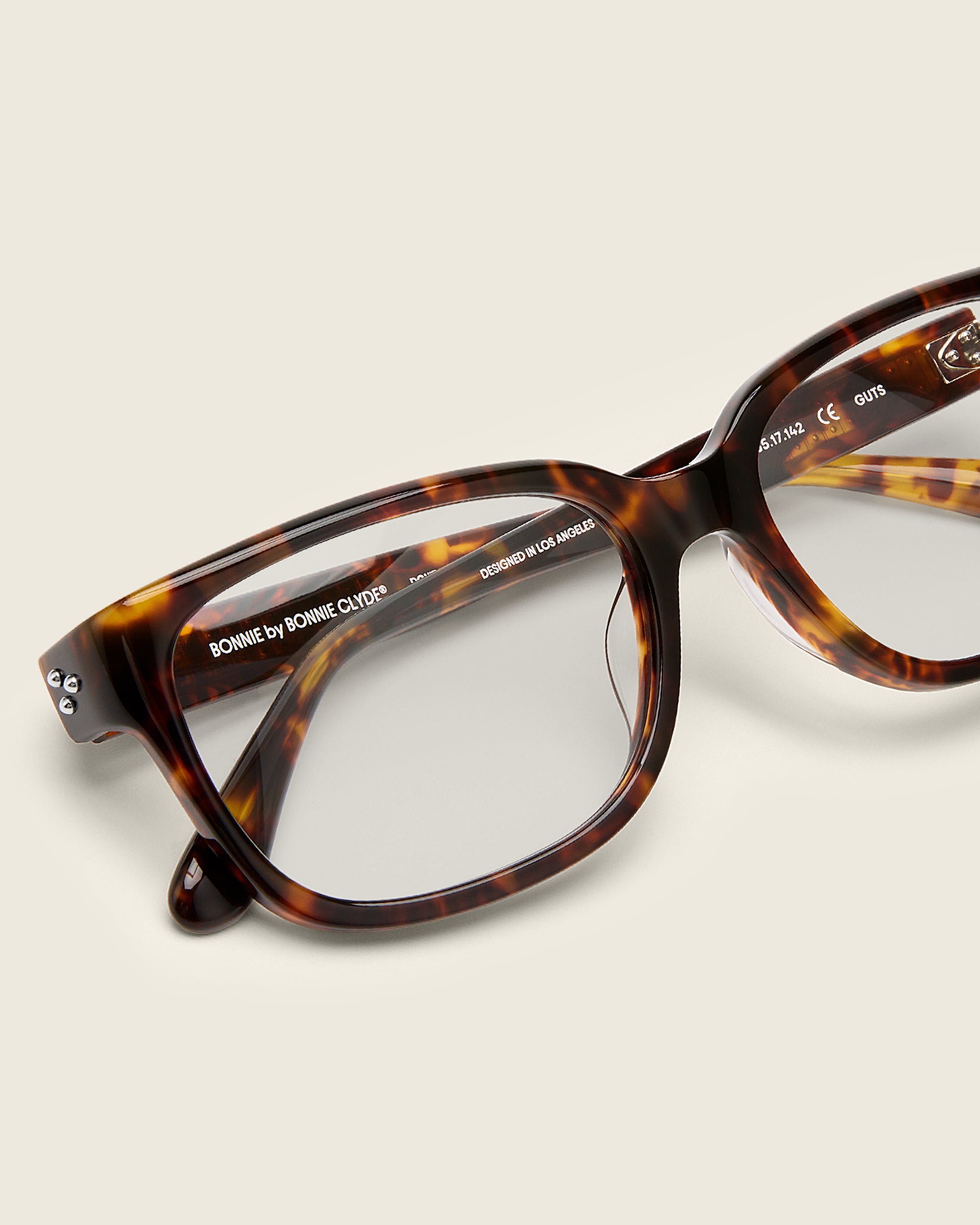 Bold, square-shaped tortoise acetate frames on a beige background. Detail view. Mirrored metal bead detail on arm temples.