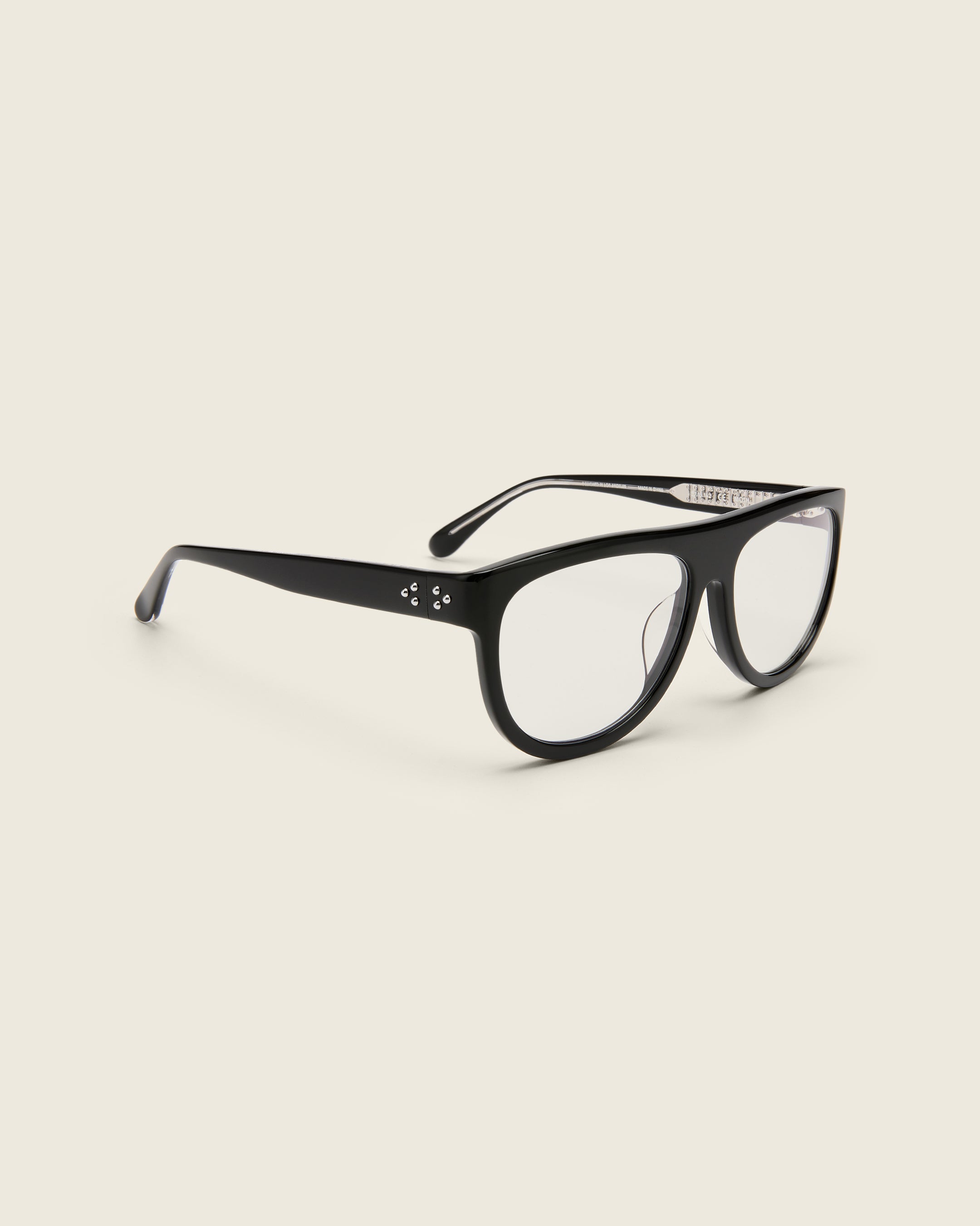 Oversized aviator-style black acetate frames with a bold browline and deep lenses on a cream background. Quarter view. Mirrored triple metal bead detailing on hinge and arm temple.