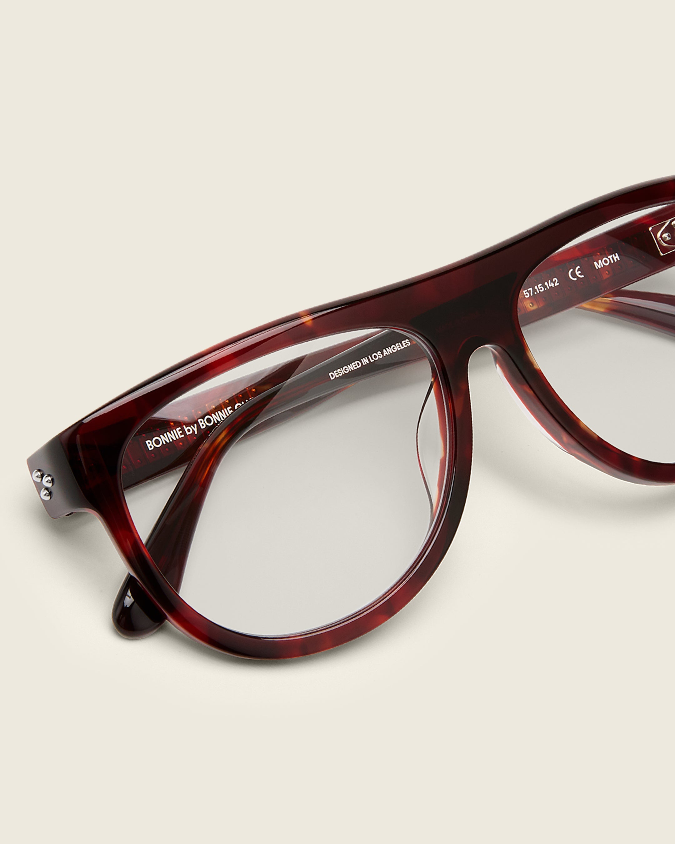 Oversized aviator-style red tortoise acetate frames with a bold browline and deep lenses on a cream background. Detail view. Mirrored triple metal bead detailing on hinge and arm temple.