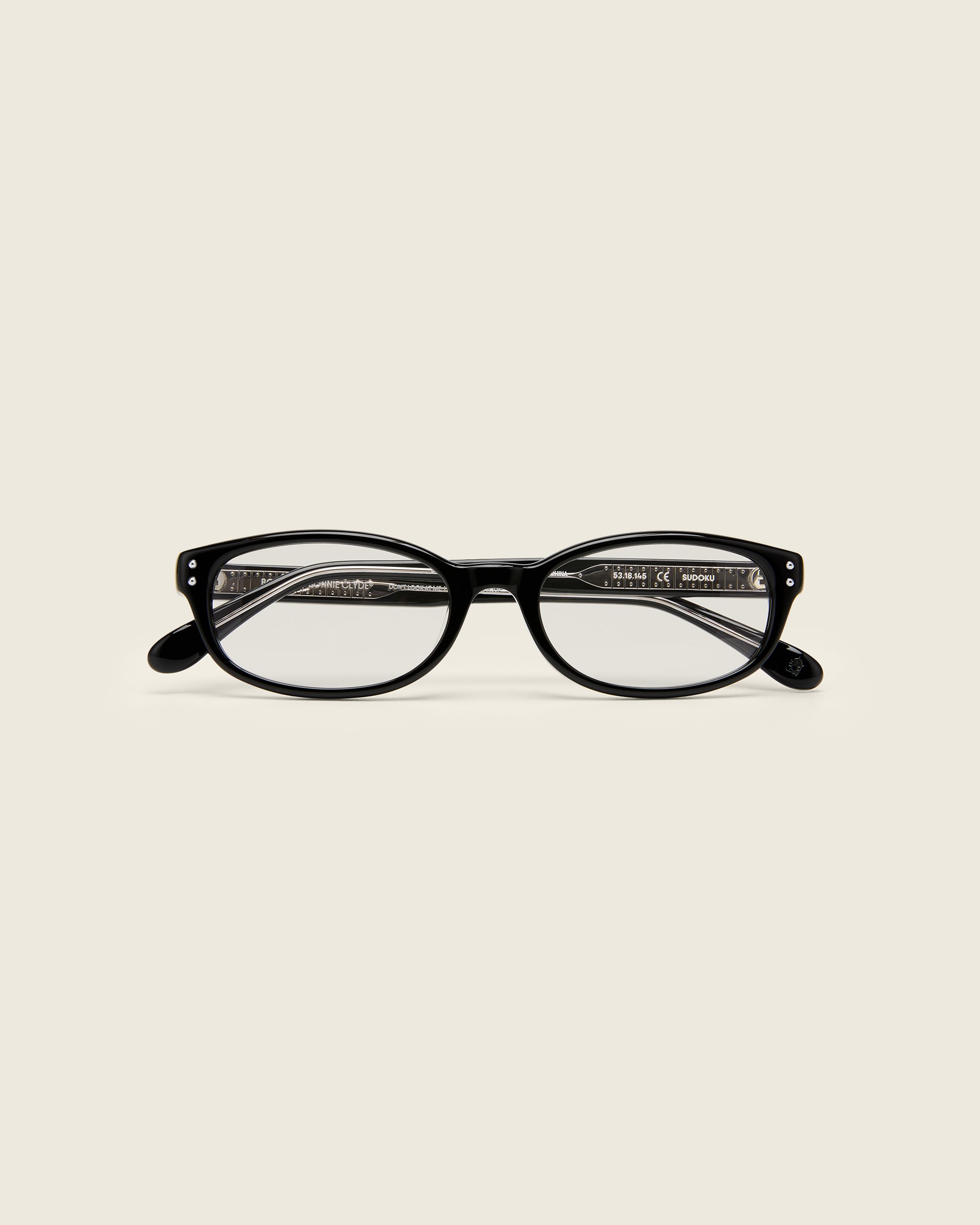 Black acetate oval frames with a curved nosebridge and extended ear tips on a cream background. Face view. Double metal detailing on ear tips.