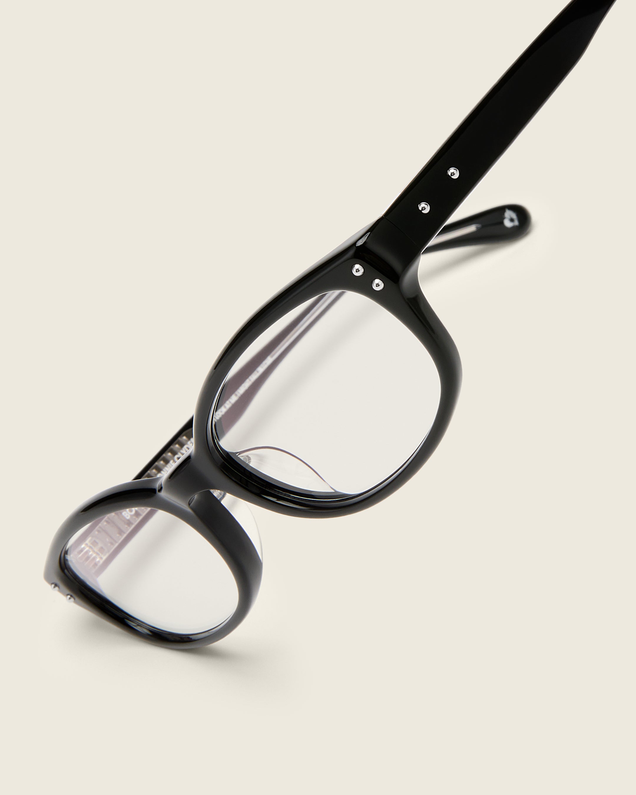 Black acetate oval frames with a curved nosebridge and extended ear tips on a cream background. Detail view. Matching double metal detailing on ear tips and arm temples.