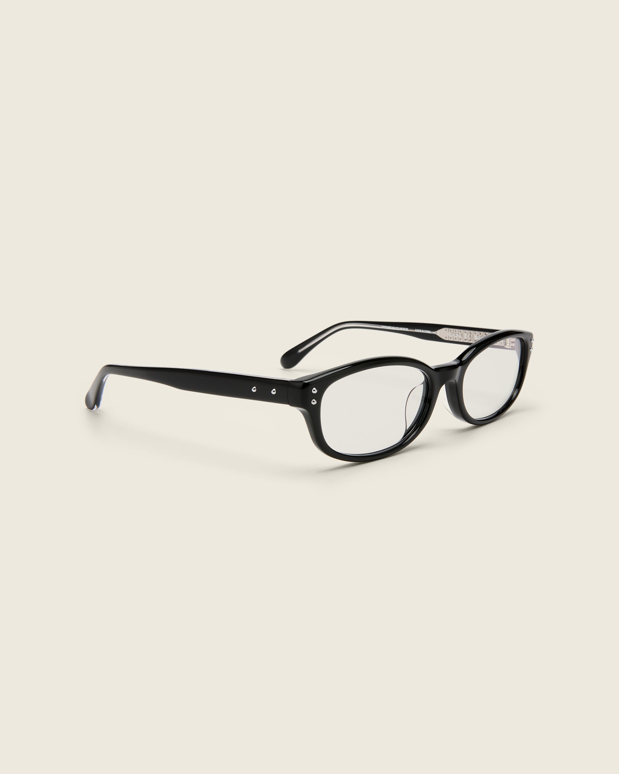 Black acetate oval frames with a curved nosebridge and extended ear tips on a cream background. Quarter view. Matching double metal detailing on ear tips and arm temples.