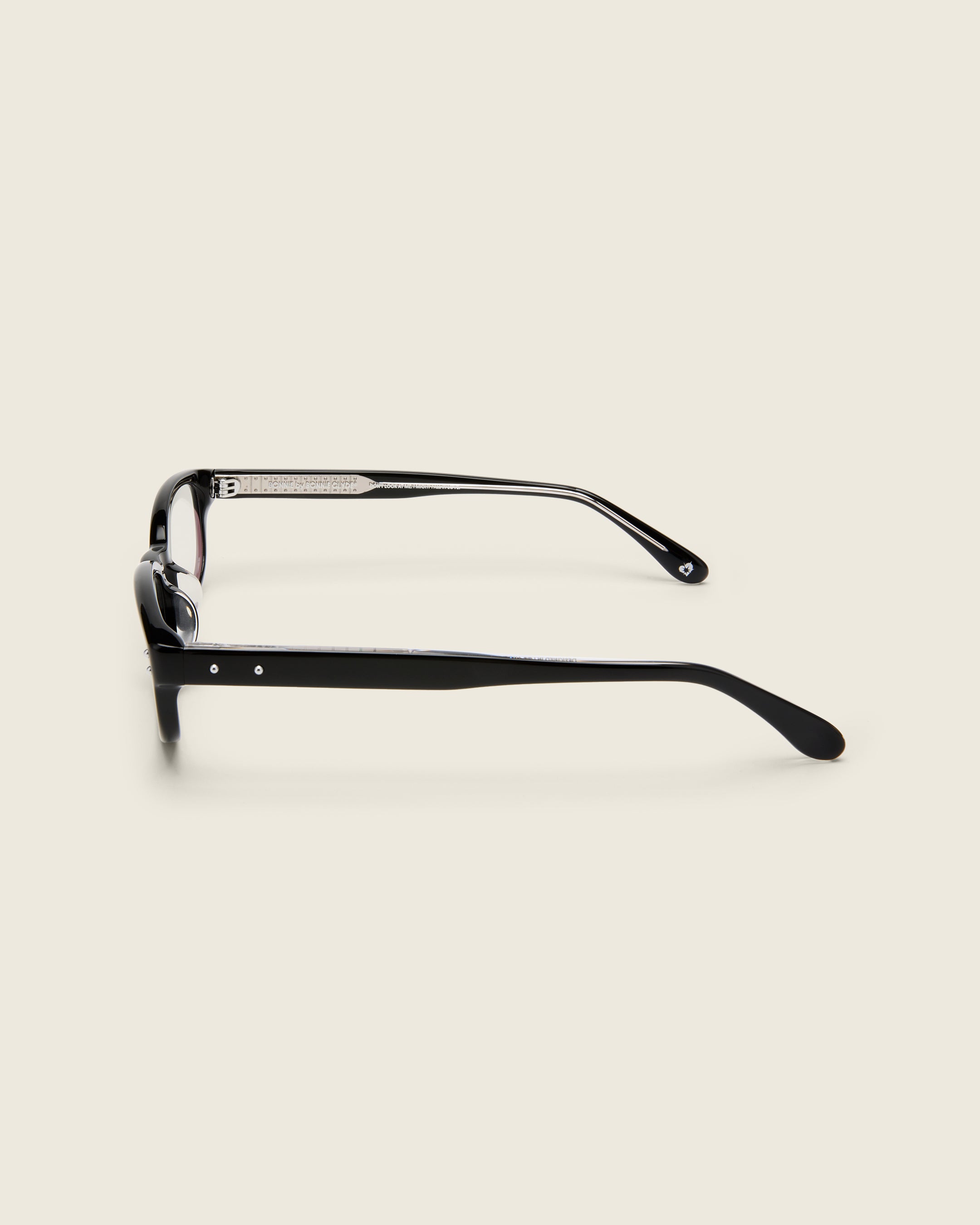 Black acetate oval frames with a curved nosebridge and extended ear tips on a cream background. Side view. Matching double metal detailing on ear tips and arm temples.
