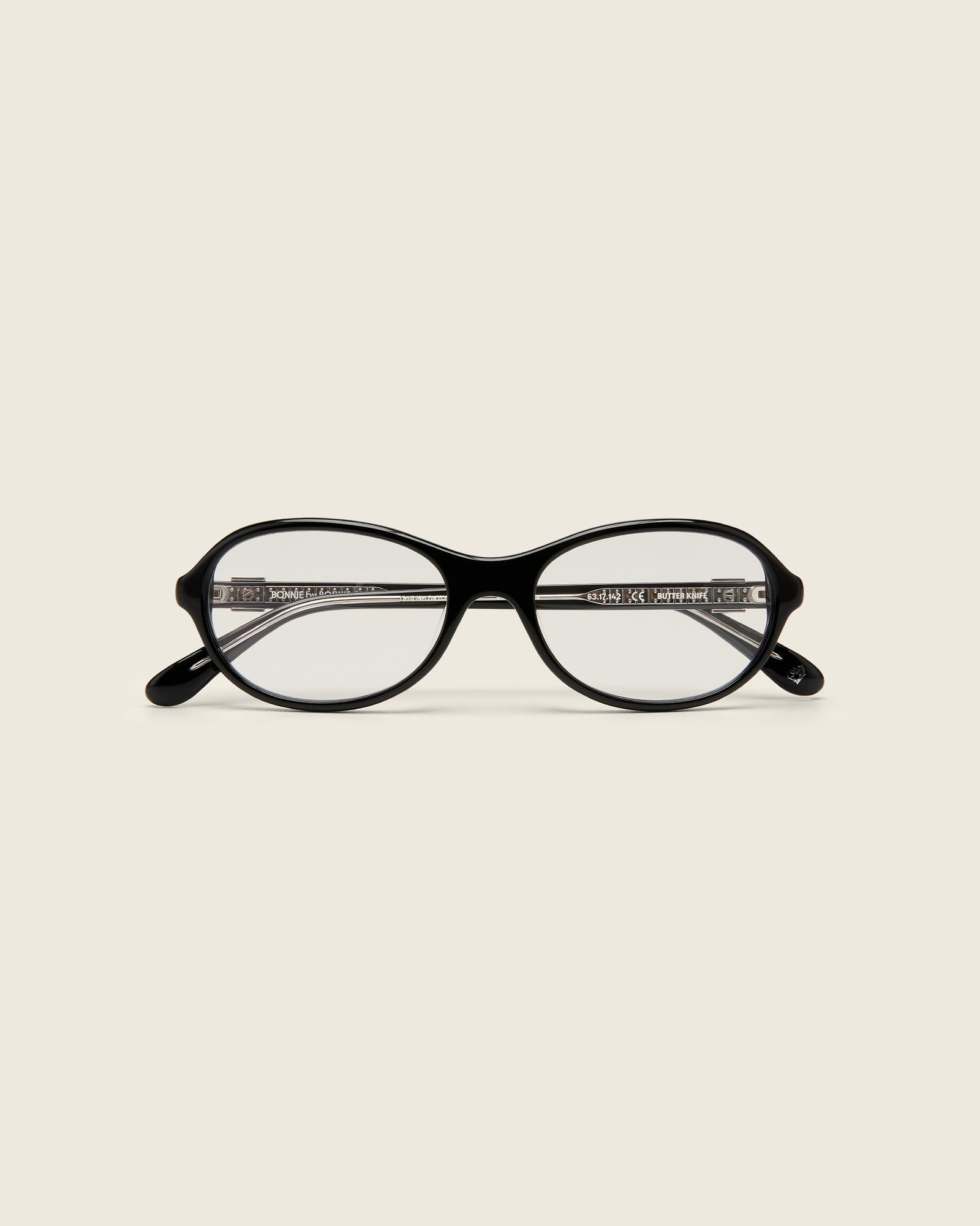 Rounded black acetate frames. Features raised browline curvature on a beige background. Features custom metal heart logo detail on arm temple. Face view.