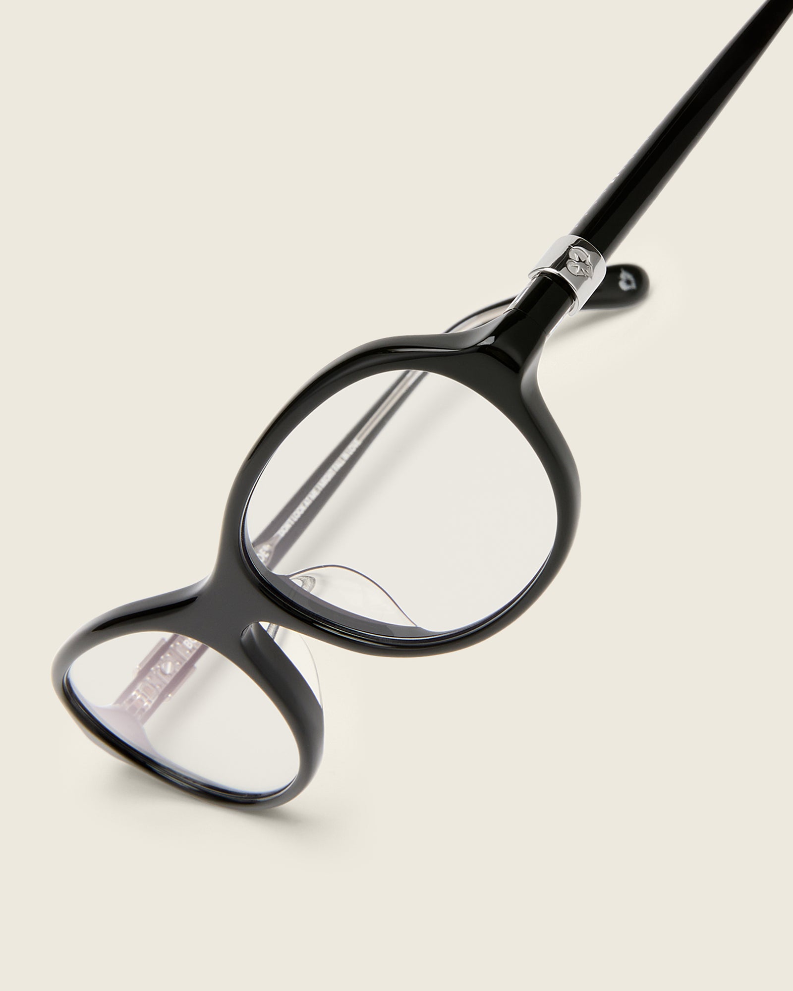 Rounded black acetate frames. Features raised browline curvature on a beige background. Features custom metal heart logo detail on arm temple. Detail view.