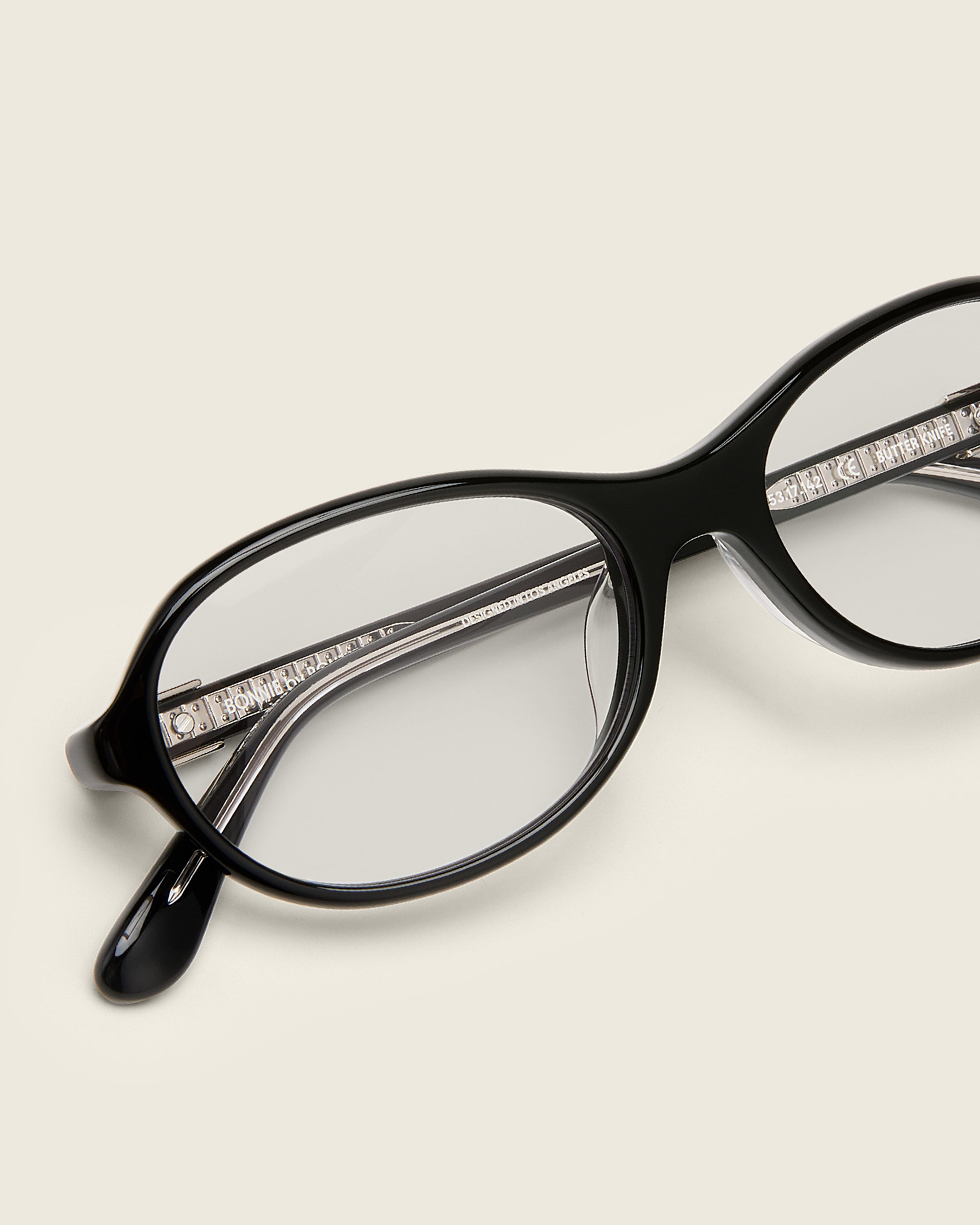 Rounded black acetate frames. Features raised browline curvature on a beige background. Features custom metal heart logo detail on arm temple. Detail view.