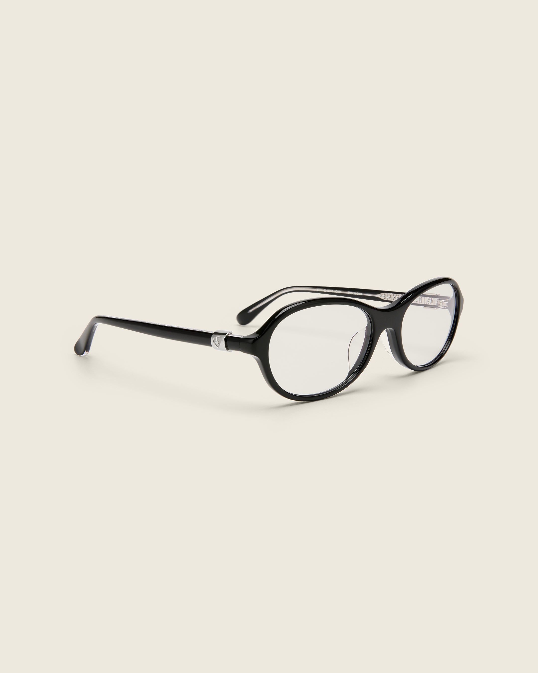 Rounded black acetate frames. Features raised browline curvature on a beige background. Features custom metal heart logo detail on arm temple. Quarter view.