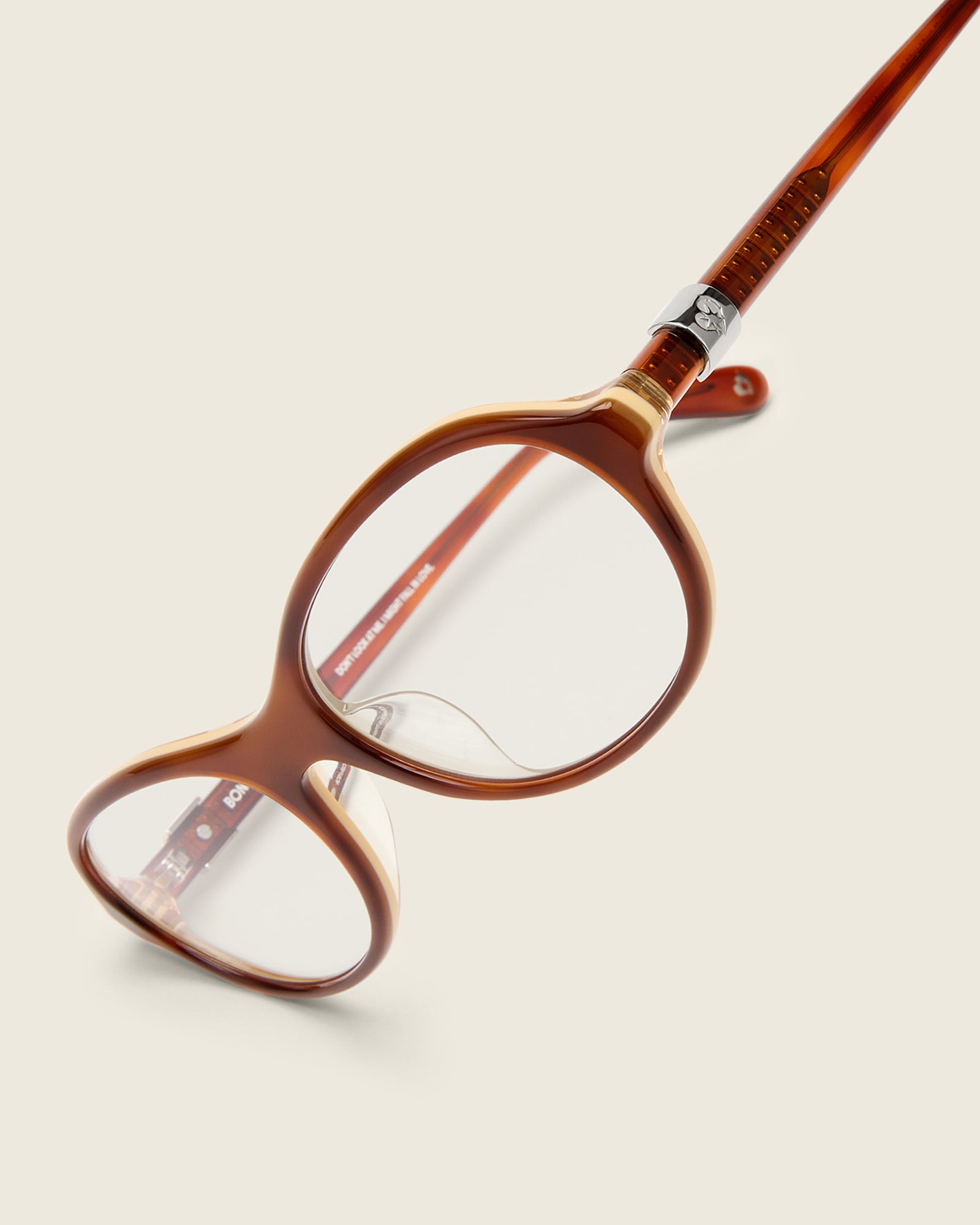 Rounded brown acetate frames. Features raised browline curvature on a beige background. Features custom metal heart logo detail on arm temple. Detail view.