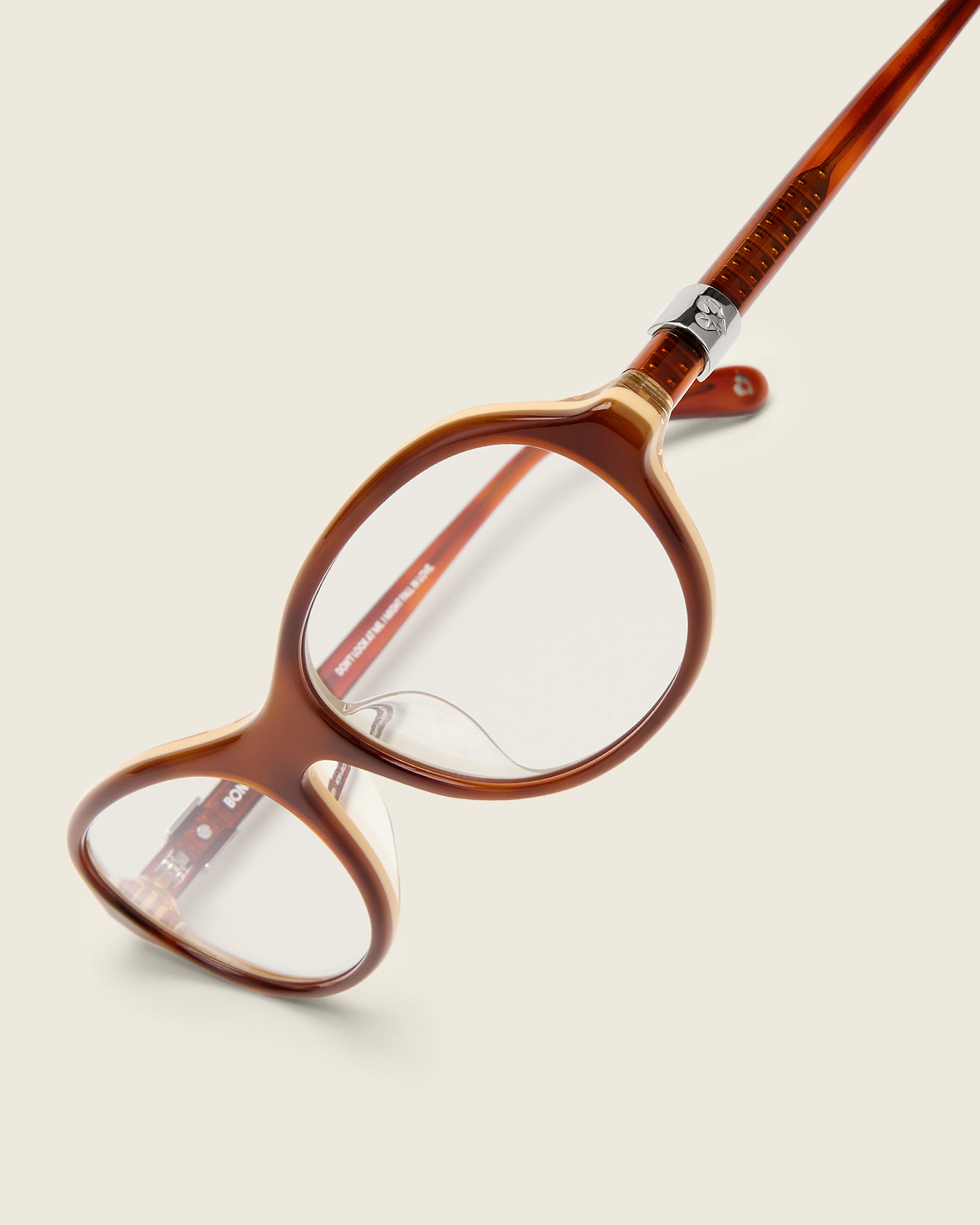 Rounded brown acetate frames. Features raised browline curvature on a beige background. Features custom metal heart logo detail on arm temple. Detail view.