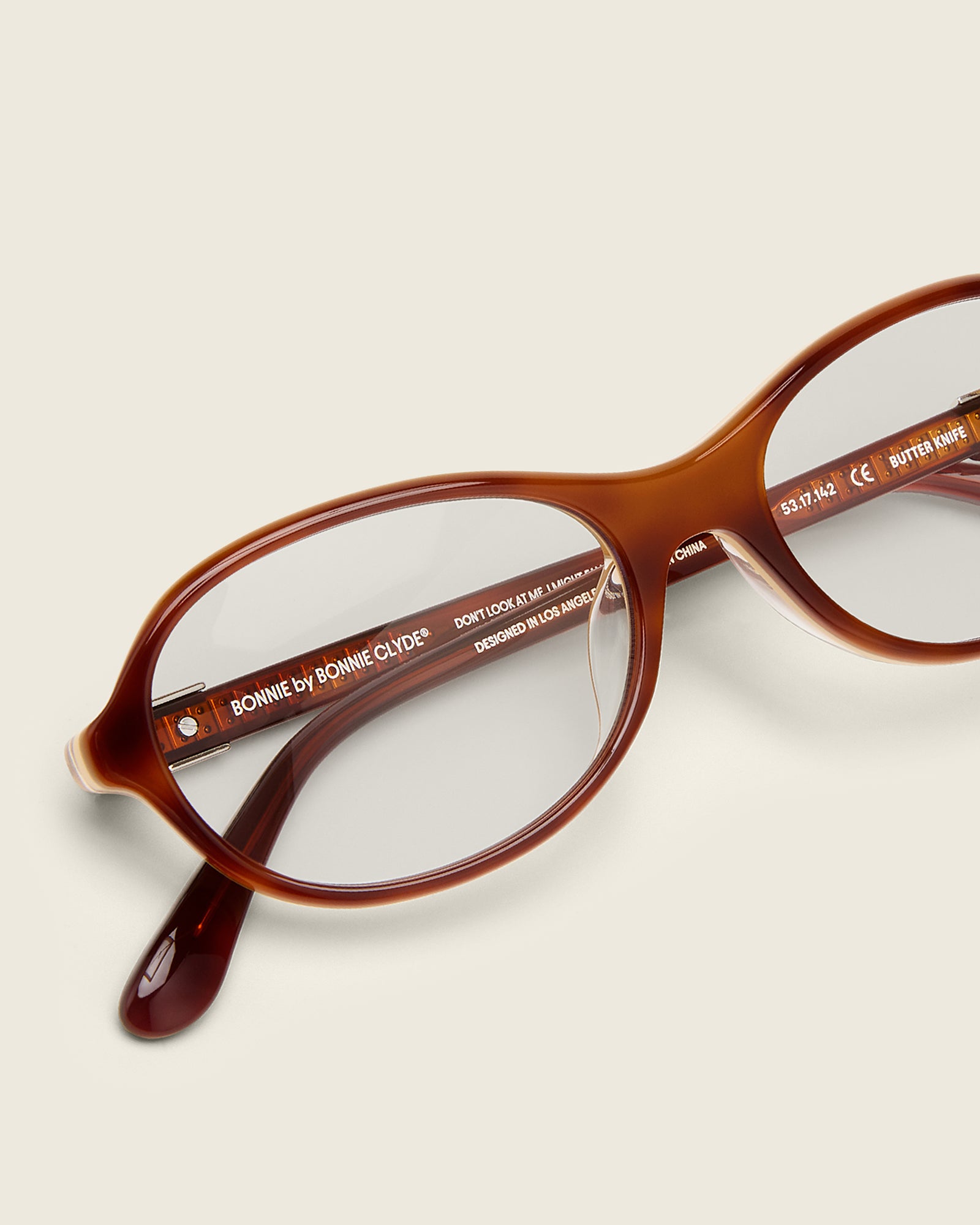 Rounded brown acetate frames. Features raised browline curvature on a beige background. Features custom metal heart logo detail on arm temple. Detail view.