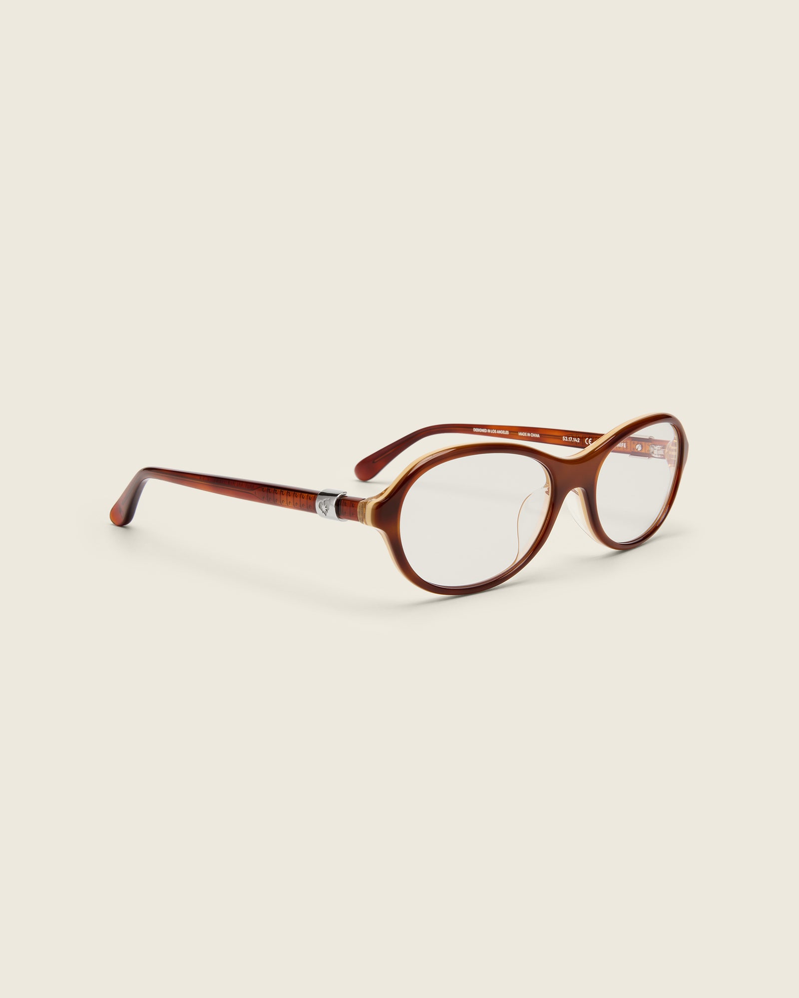 Rounded brown acetate frames. Features raised browline curvature on a beige background. Features custom metal heart logo detail on arm temple. Quarter view.