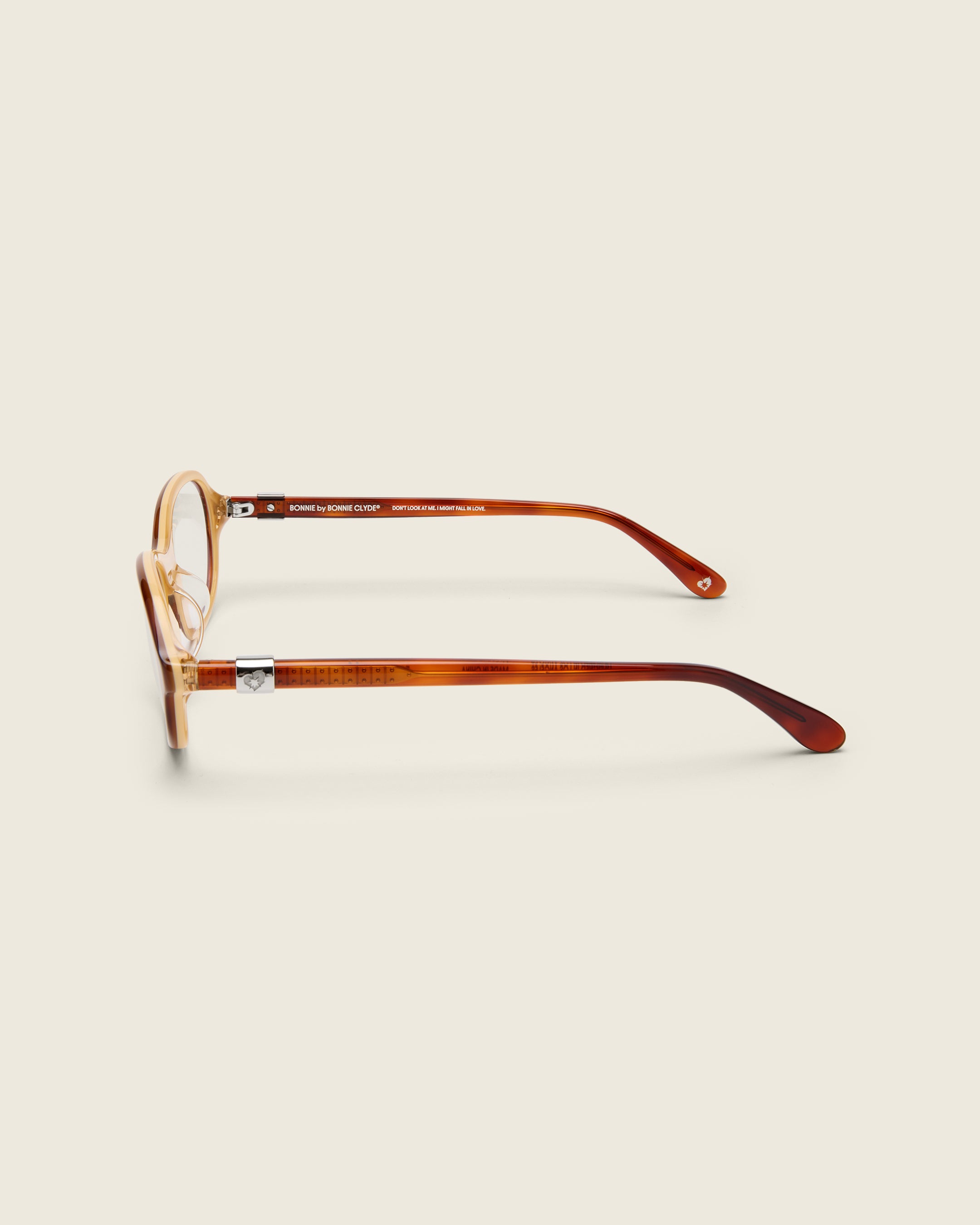 Rounded brown acetate frames. Features raised browline curvature on a beige background. Features custom metal heart logo detail on arm temple. Side view.
