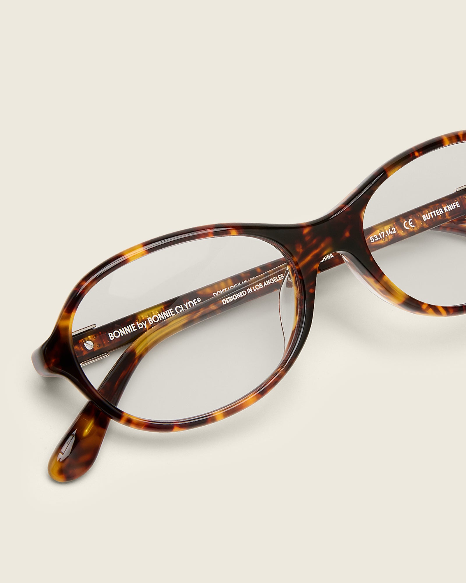 Rounded tortoise acetate frames. Features raised browline curvature on a beige background. Features custom metal heart logo detail on arm temple. Detail view.