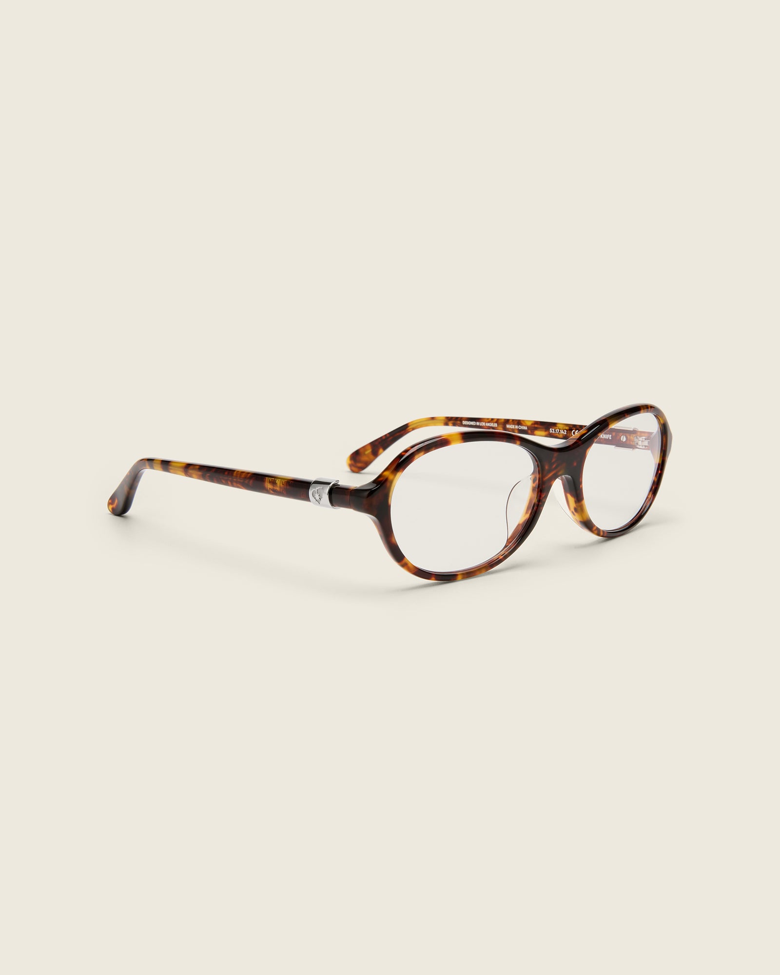 Rounded tortoise acetate frames. Features raised browline curvature on a beige background. Features custom metal heart logo detail on arm temple. Quarter view.