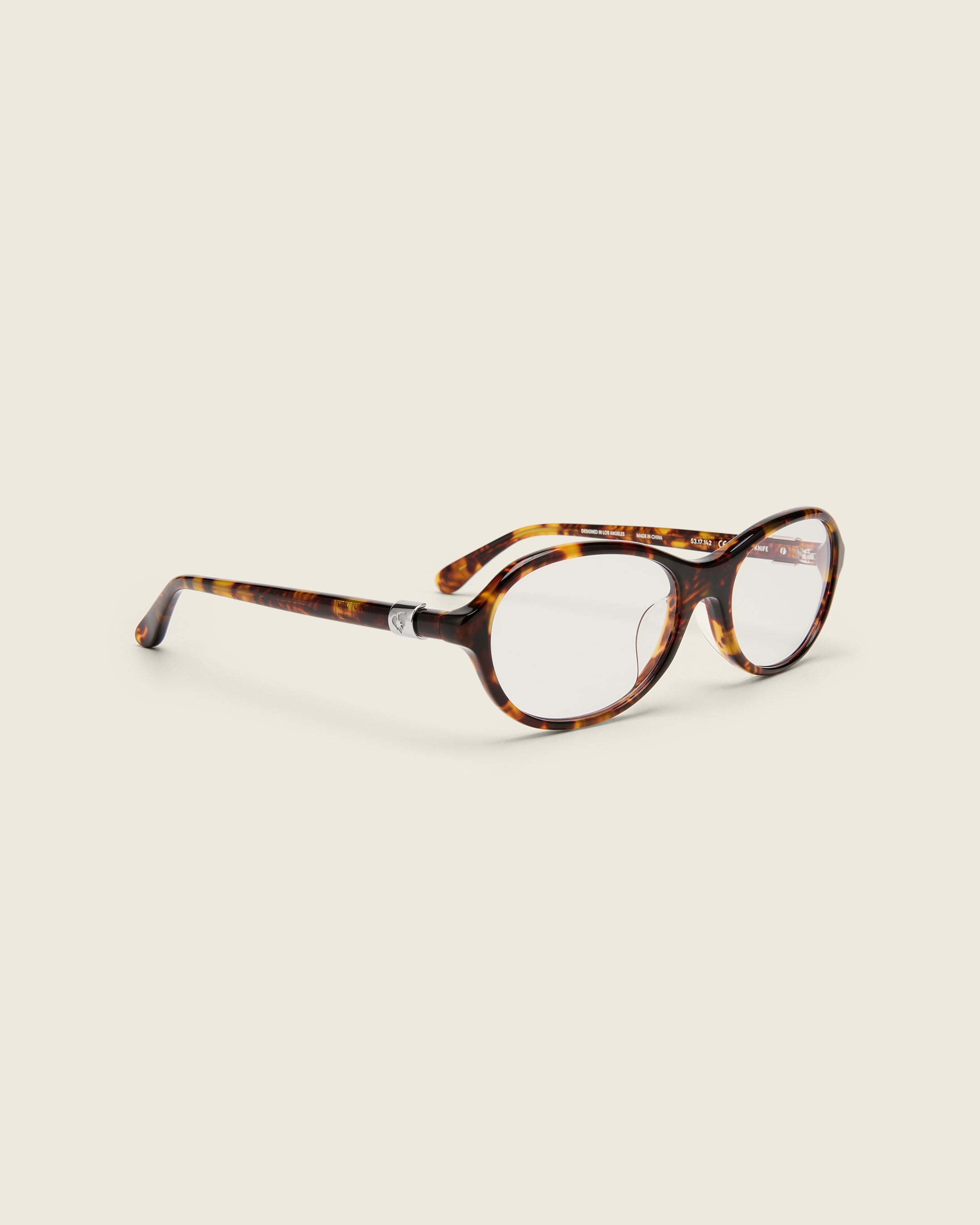 Rounded tortoise acetate frames. Features raised browline curvature on a beige background. Features custom metal heart logo detail on arm temple. Quarter view.