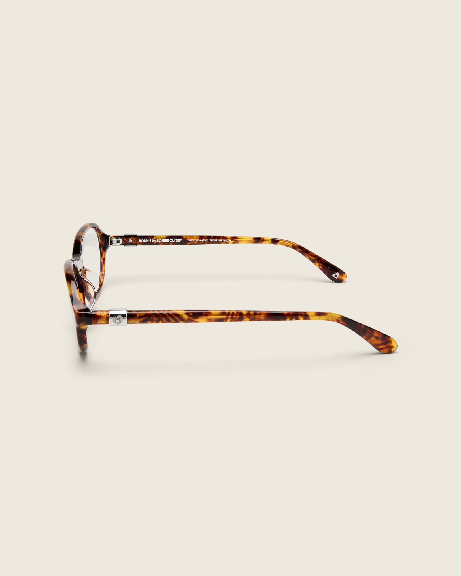 Rounded tortoise acetate frames. Features raised browline curvature on a beige background. Features custom metal heart logo detail on arm temple. Side view.