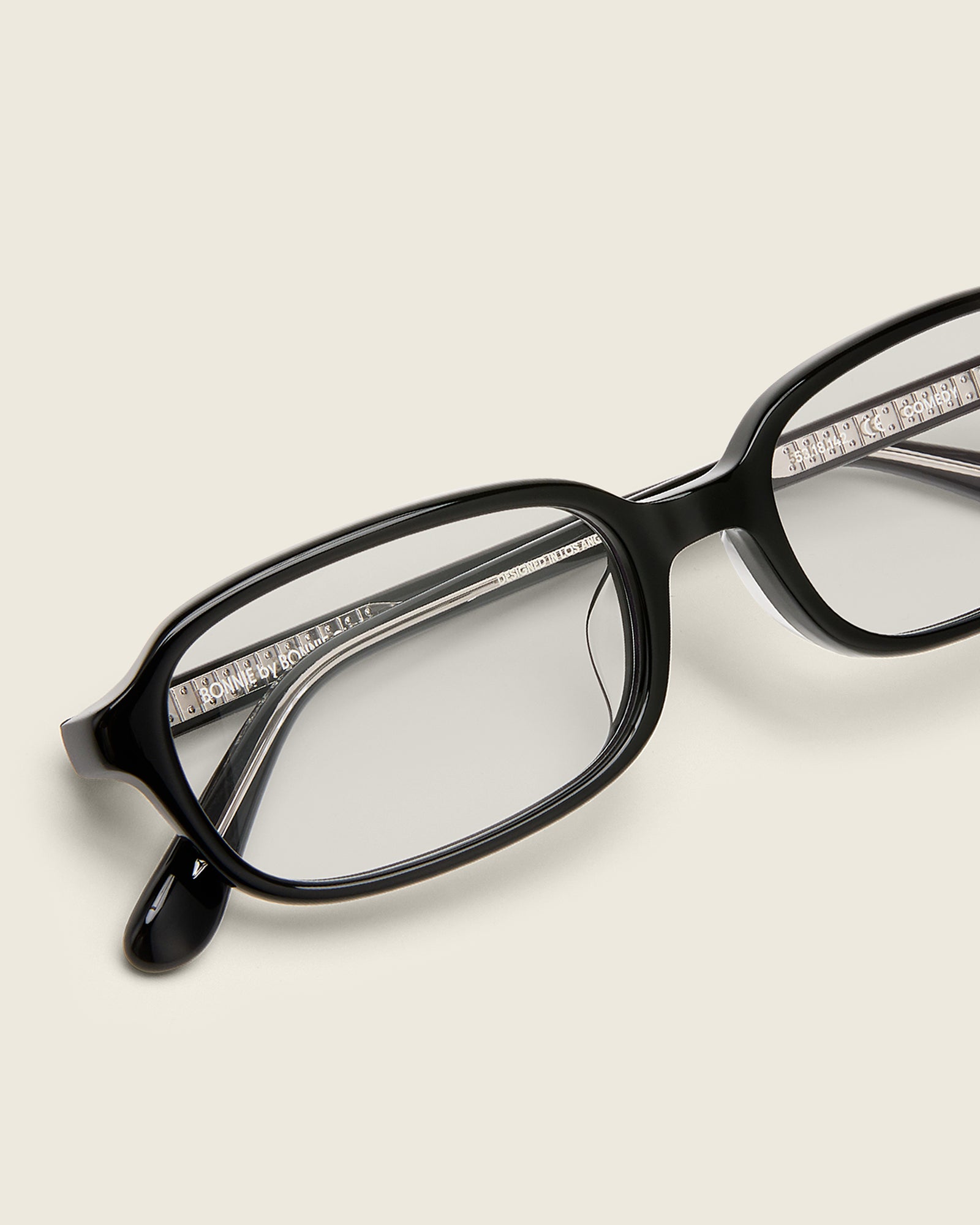 Rectangular black acetate frames with softened corners and a low-profile bridge on a beige background. Detail view.