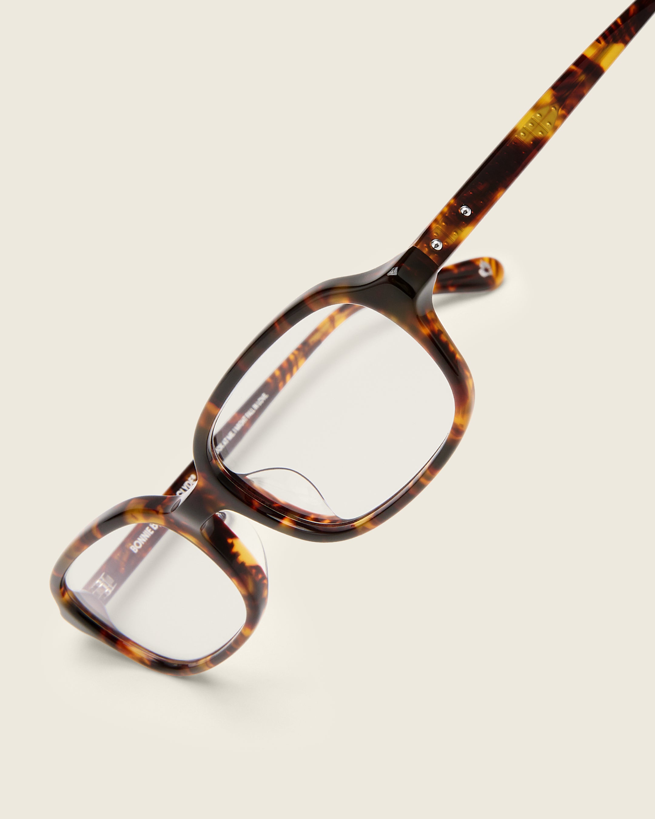 Rectangular tortoise acetate frames with softened corners and a low-profile bridge on a beige background. Detail view.