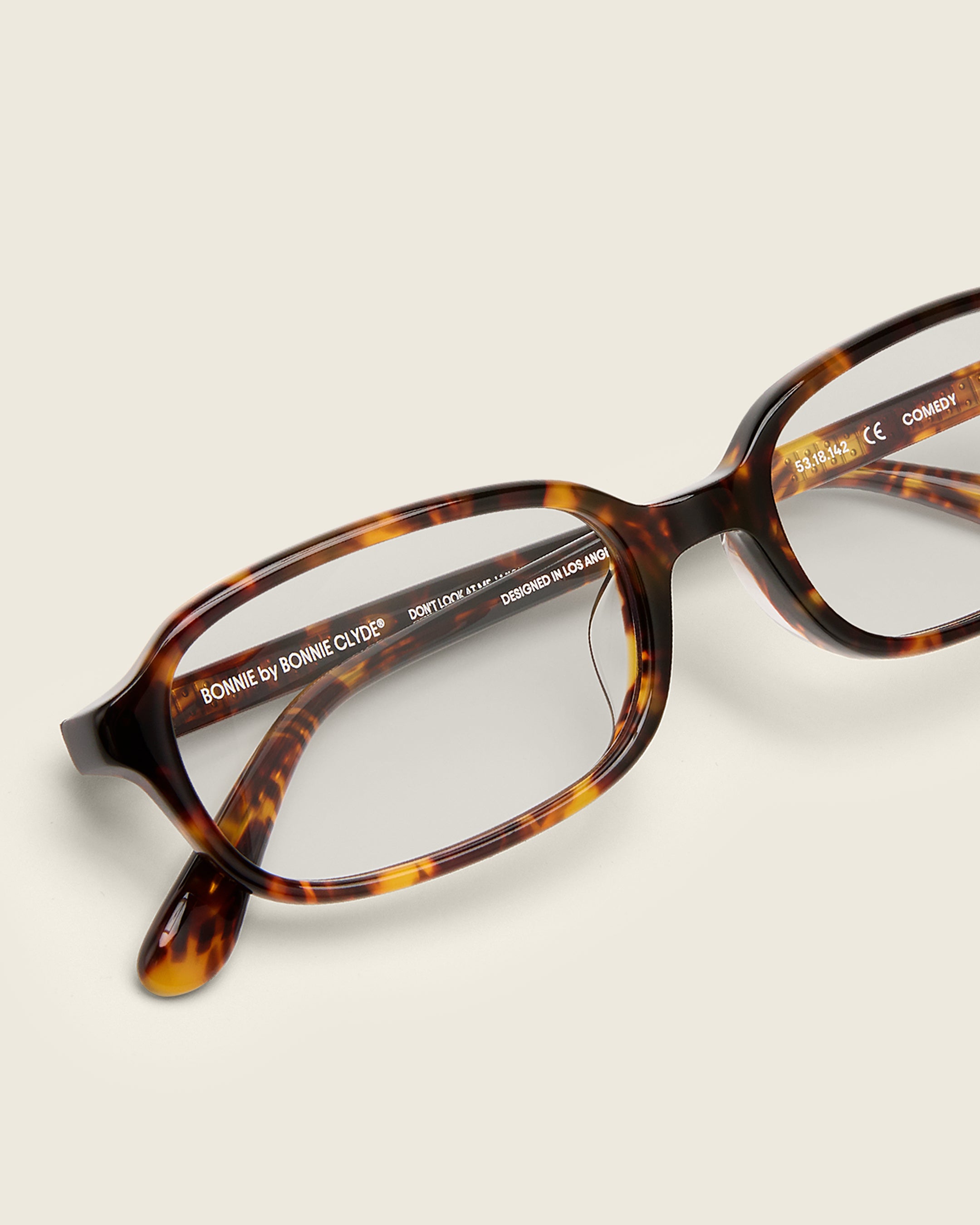 Rectangular tortoise acetate frames with softened corners and a low-profile bridge on a beige background. Detail view.