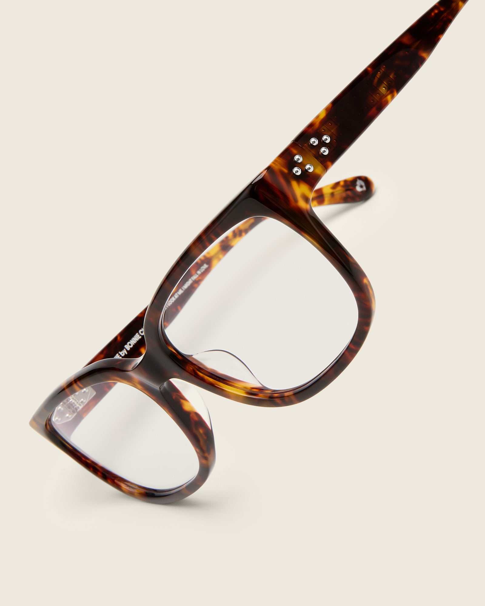 Bold, square-shaped tortoise acetate frames on a beige background. Detail view. Mirrored metal bead detail on arm temples.