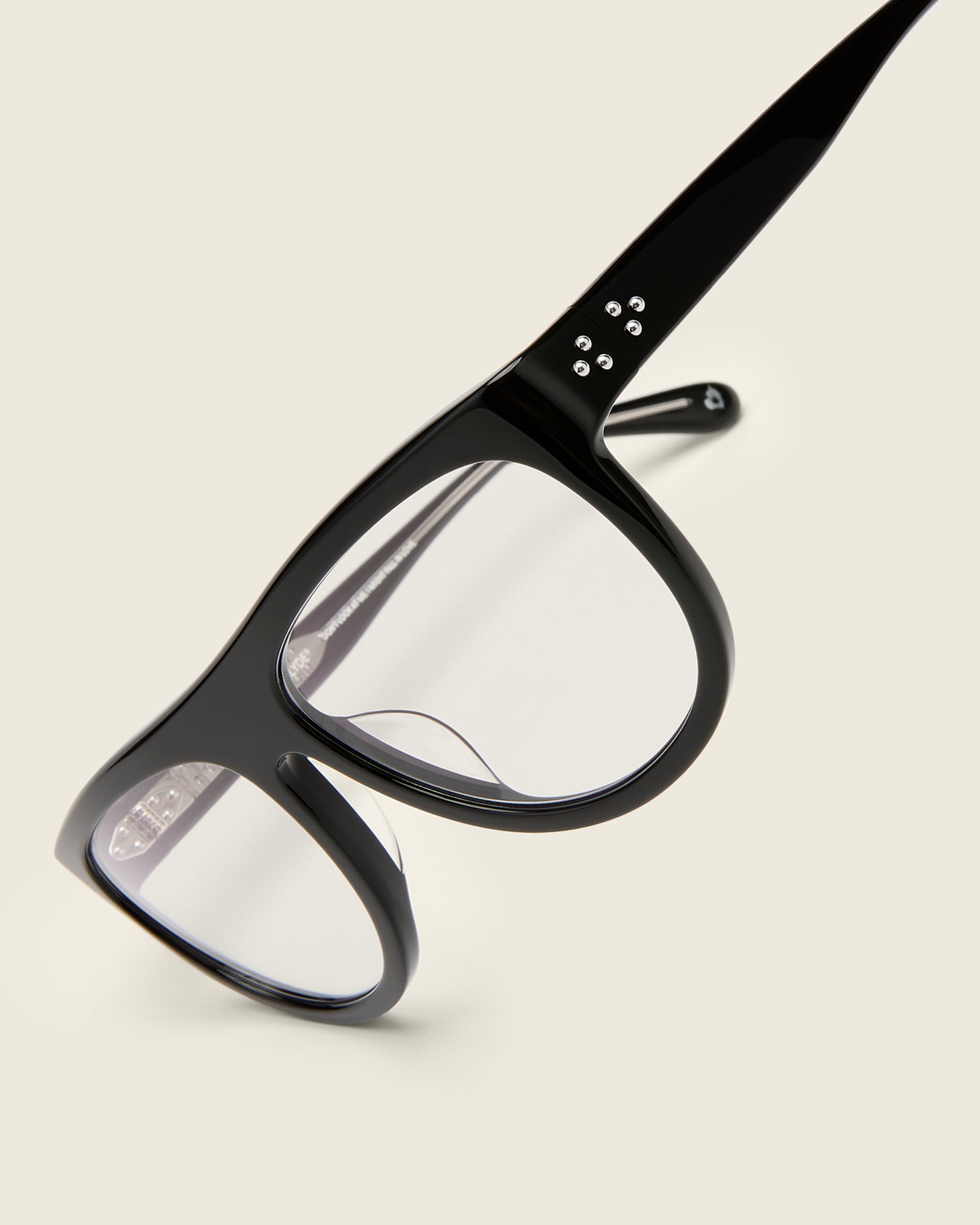 Oversized aviator-style black acetate frames with a bold browline and deep lenses on a cream background. Detail view. Mirrored triple metal bead detailing on hinge and arm temple.
