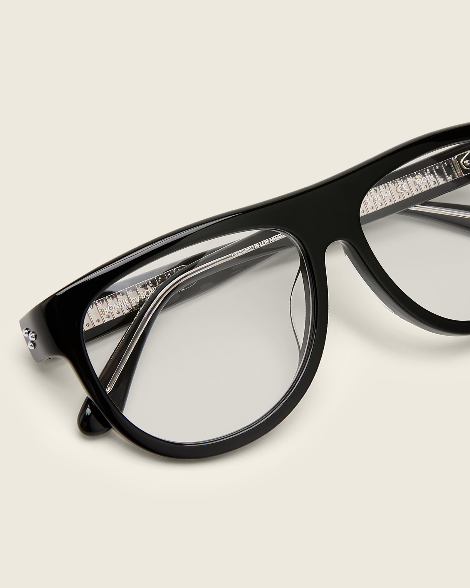 Oversized aviator-style black acetate frames with a bold browline and deep lenses on a cream background. Detail view. Mirrored triple metal bead detailing on hinge and arm temple.