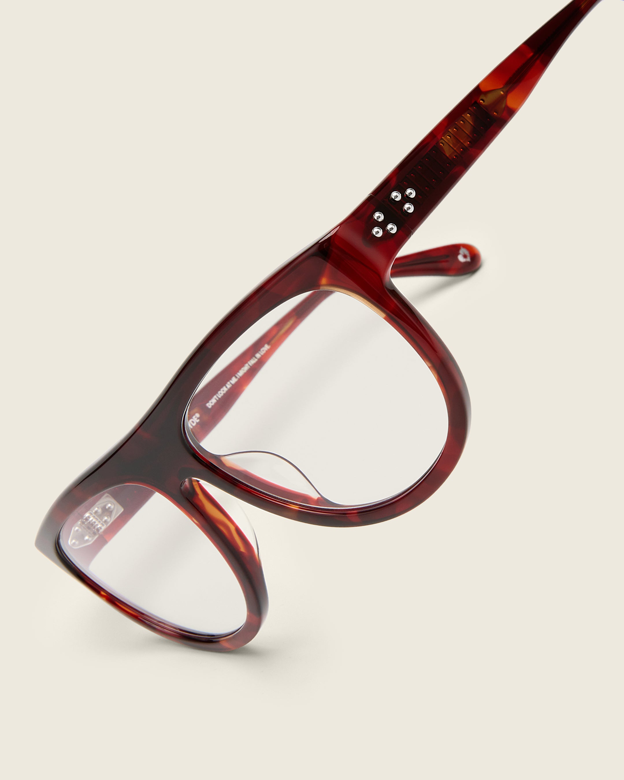 Oversized aviator-style red tortoise acetate frames with a bold browline and deep lenses on a cream background. Detail view. Mirrored triple metal bead detailing on hinge and arm temple.