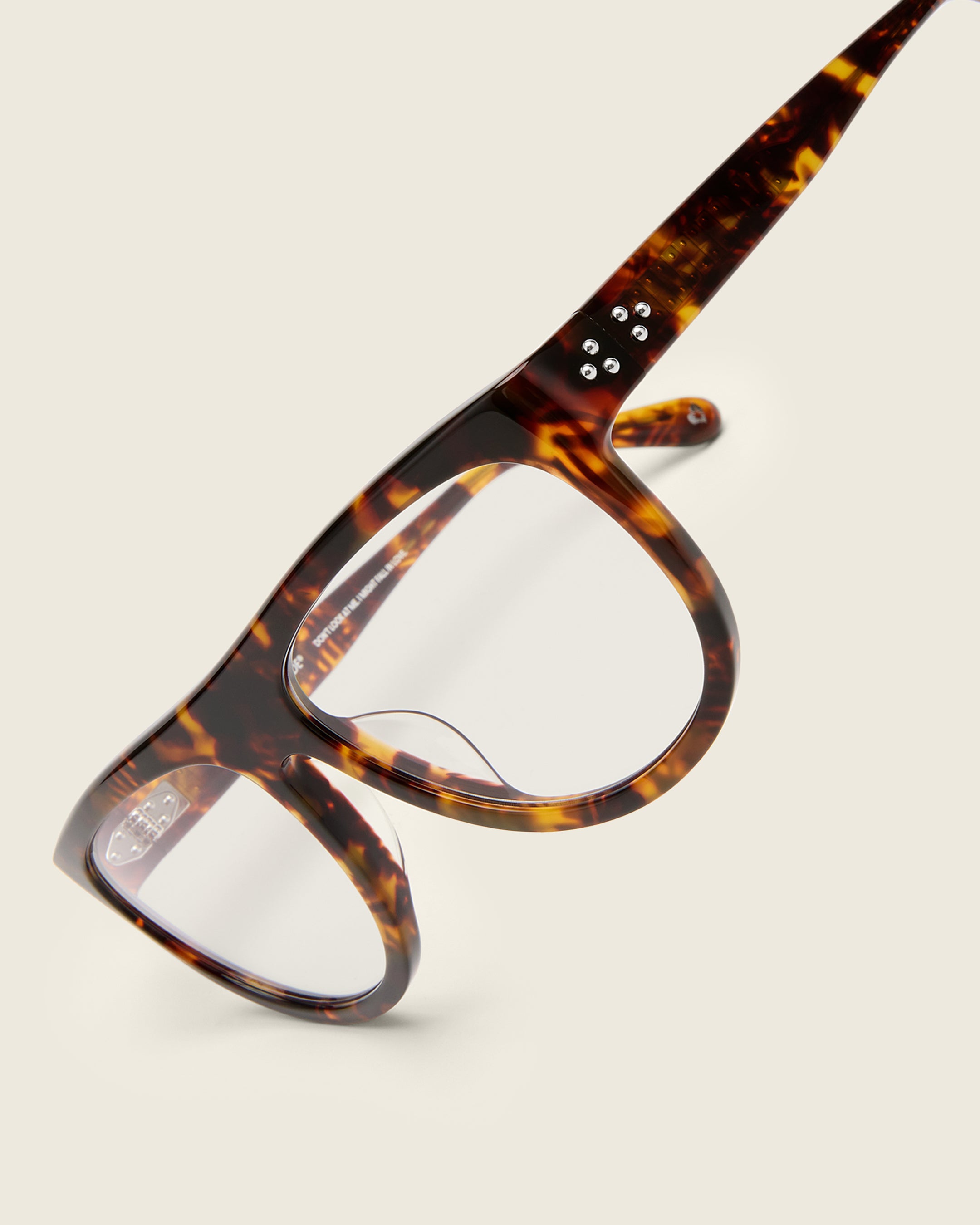 Oversized aviator-style tortoise acetate frames with a bold browline and deep lenses on a cream background. Detail view. Mirrored triple metal bead detailing on hinge and arm temple.