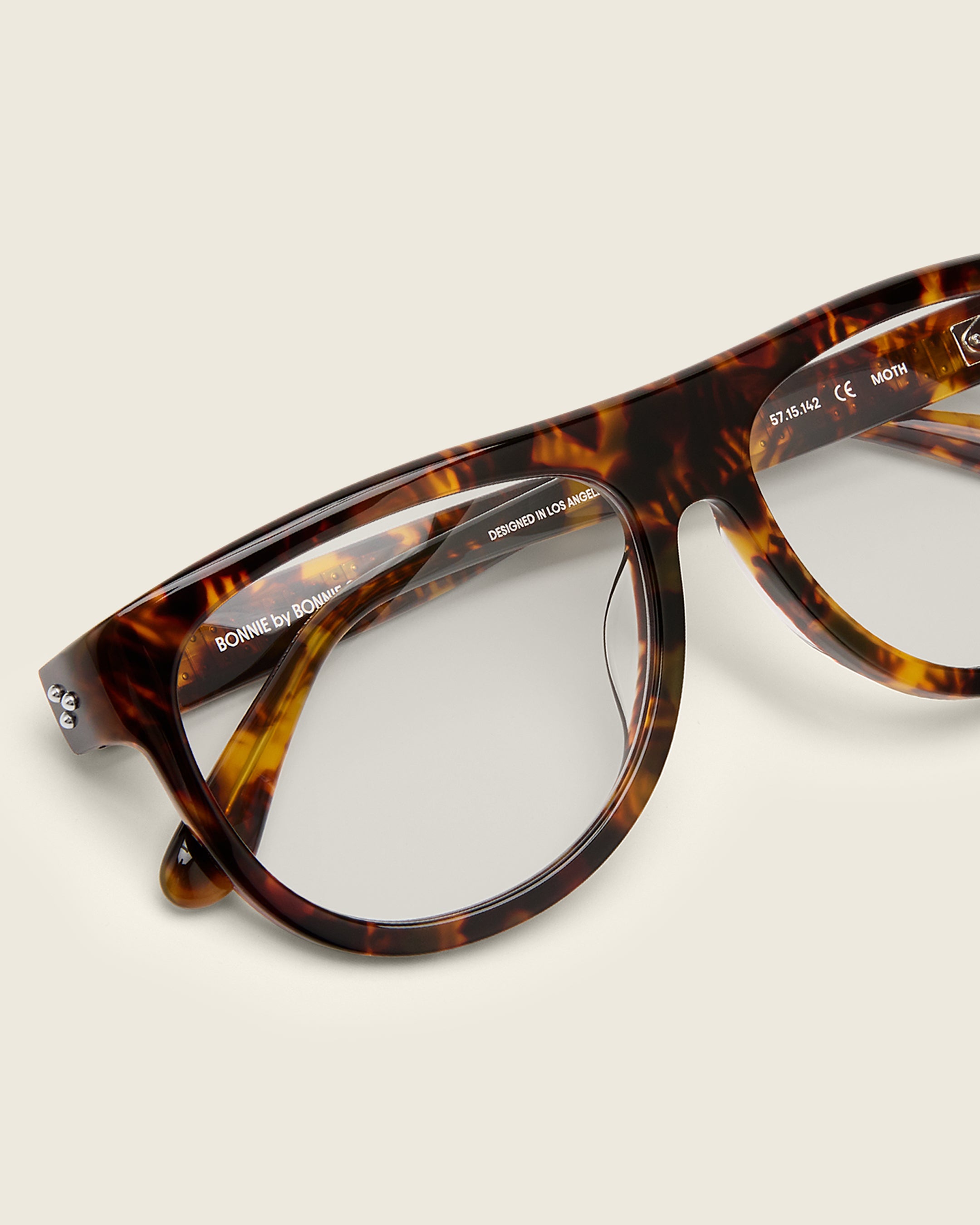 Oversized aviator-style tortoise acetate frames with a bold browline and deep lenses on a cream background. Detail view. Mirrored triple metal bead detailing on hinge and arm temple.