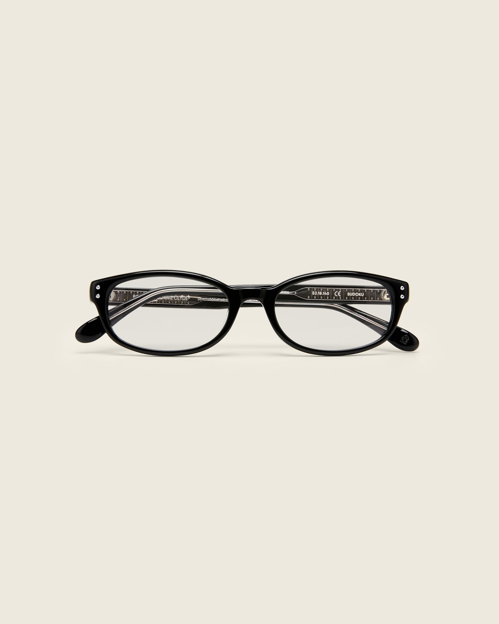 Black acetate oval frames with a curved nosebridge and extended ear tips on a cream background. Face view. Double metal detailing on ear tips.