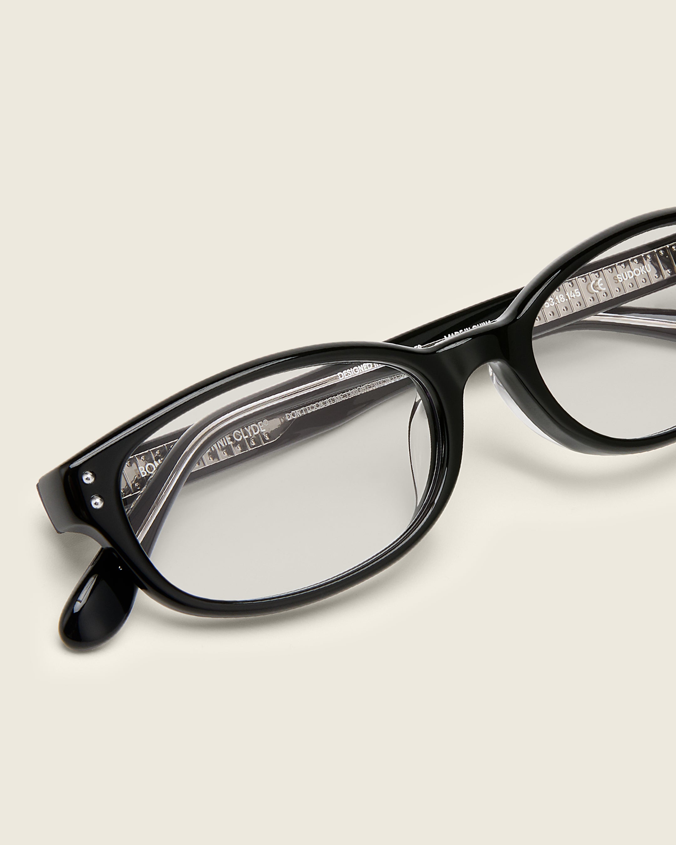 Black acetate oval frames with a curved nosebridge and extended ear tips on a cream background. Detail view. Double metal detailing on ear tips and arm temples.