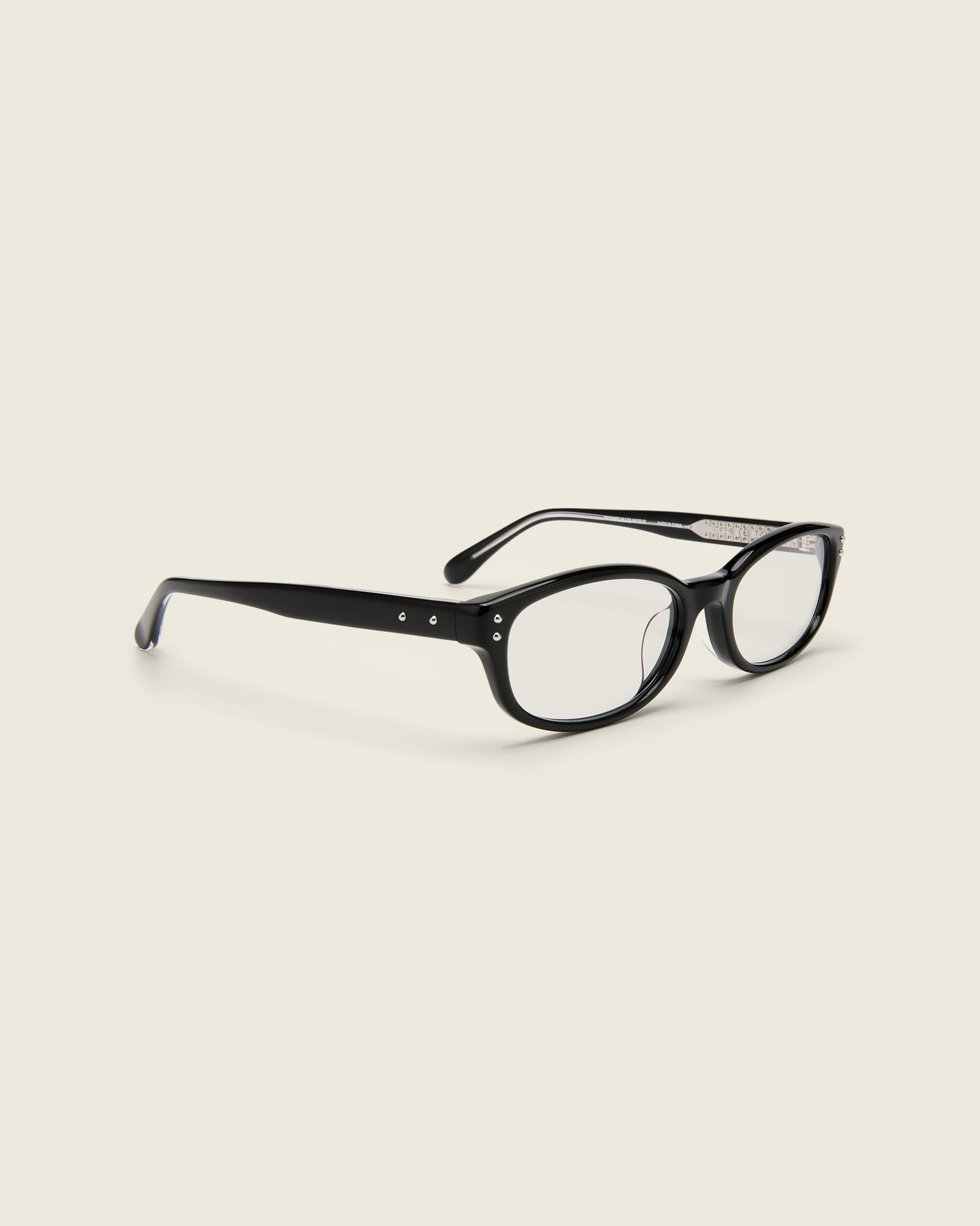 Black acetate oval frames with a curved nosebridge and extended ear tips on a cream background. Quarter view. Matching double metal detailing on ear tips and arm temples.
