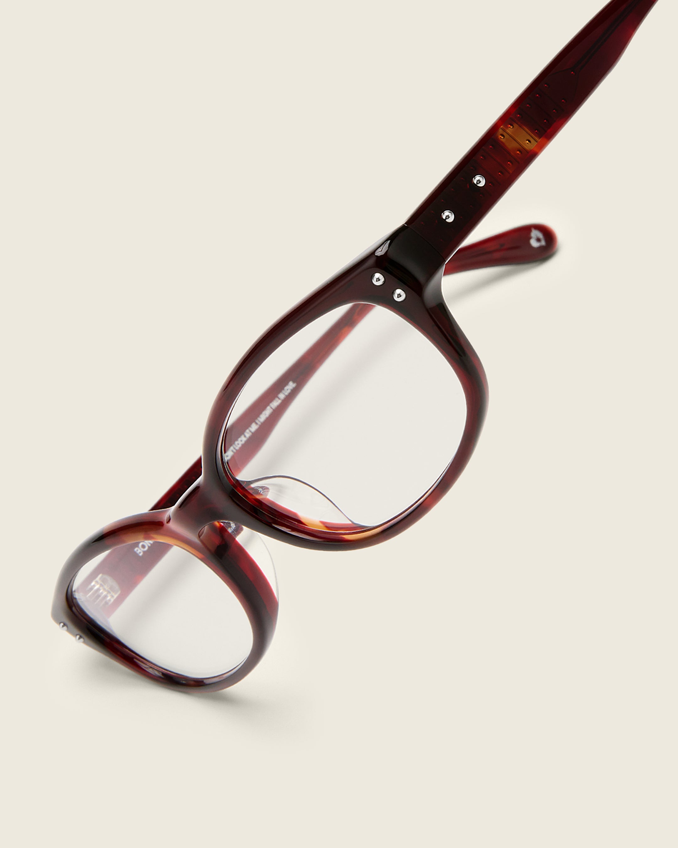 Red tortoise acetate oval frames with a curved nosebridge and extended ear tips on a cream background. Detail view. Double metal detailing on ear tips and arm temples.