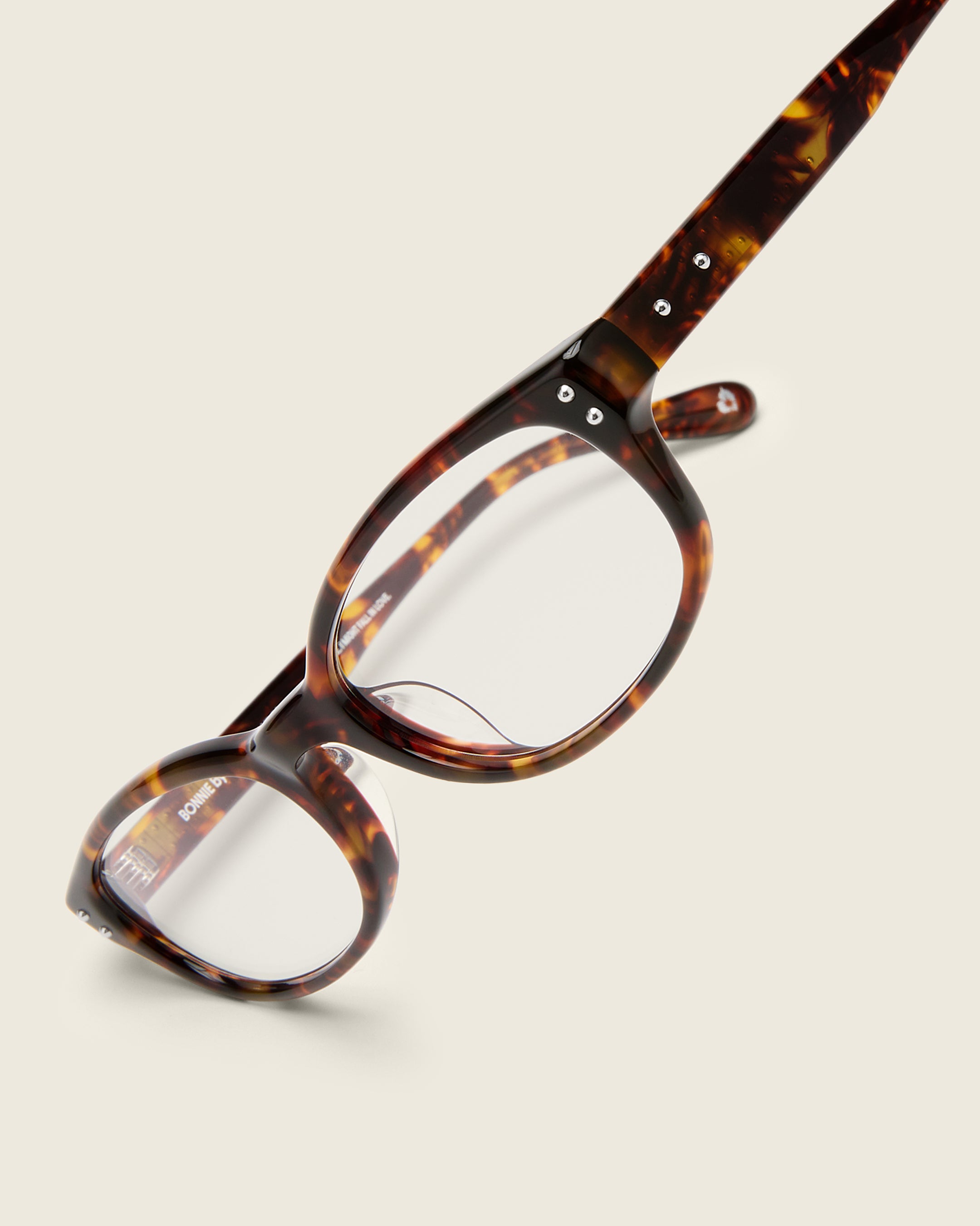 Tortoise acetate oval frames with a curved nosebridge and extended ear tips on a cream background. Detail view. Double metal detailing on ear tips and arm temples.
