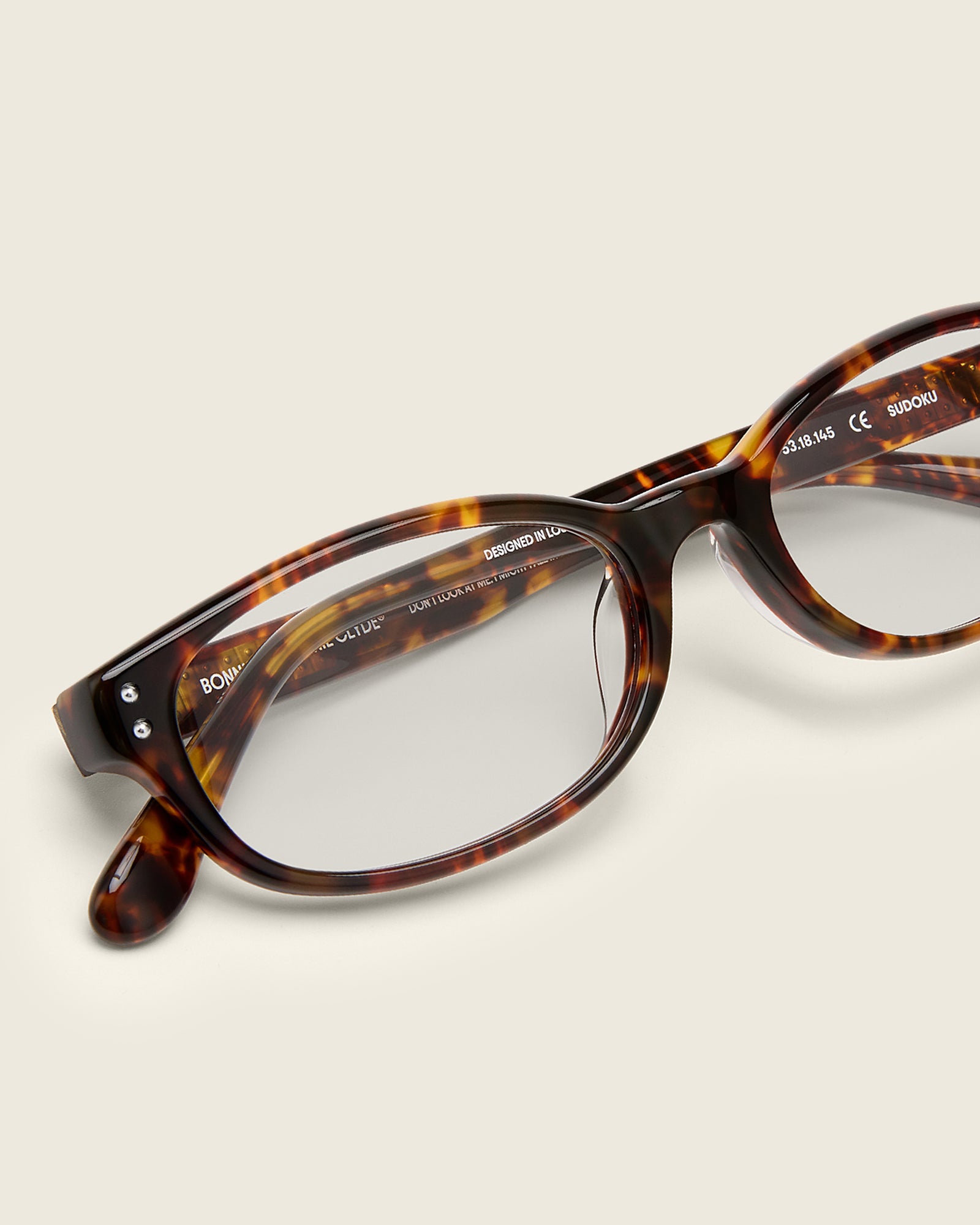 Tortoise acetate oval frames with a curved nosebridge and extended ear tips on a cream background. Detail view. Double metal detailing on ear tips and arm temples.