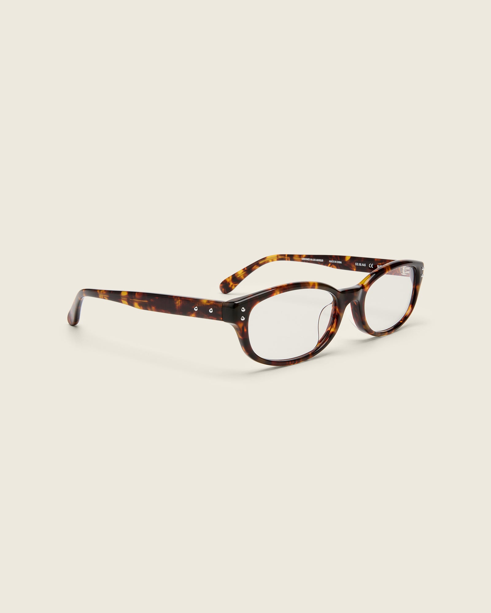 Tortoise acetate oval frames with a curved nosebridge and extended ear tips on a cream background. Quarter view. Double metal detailing on ear tips and arm temples.